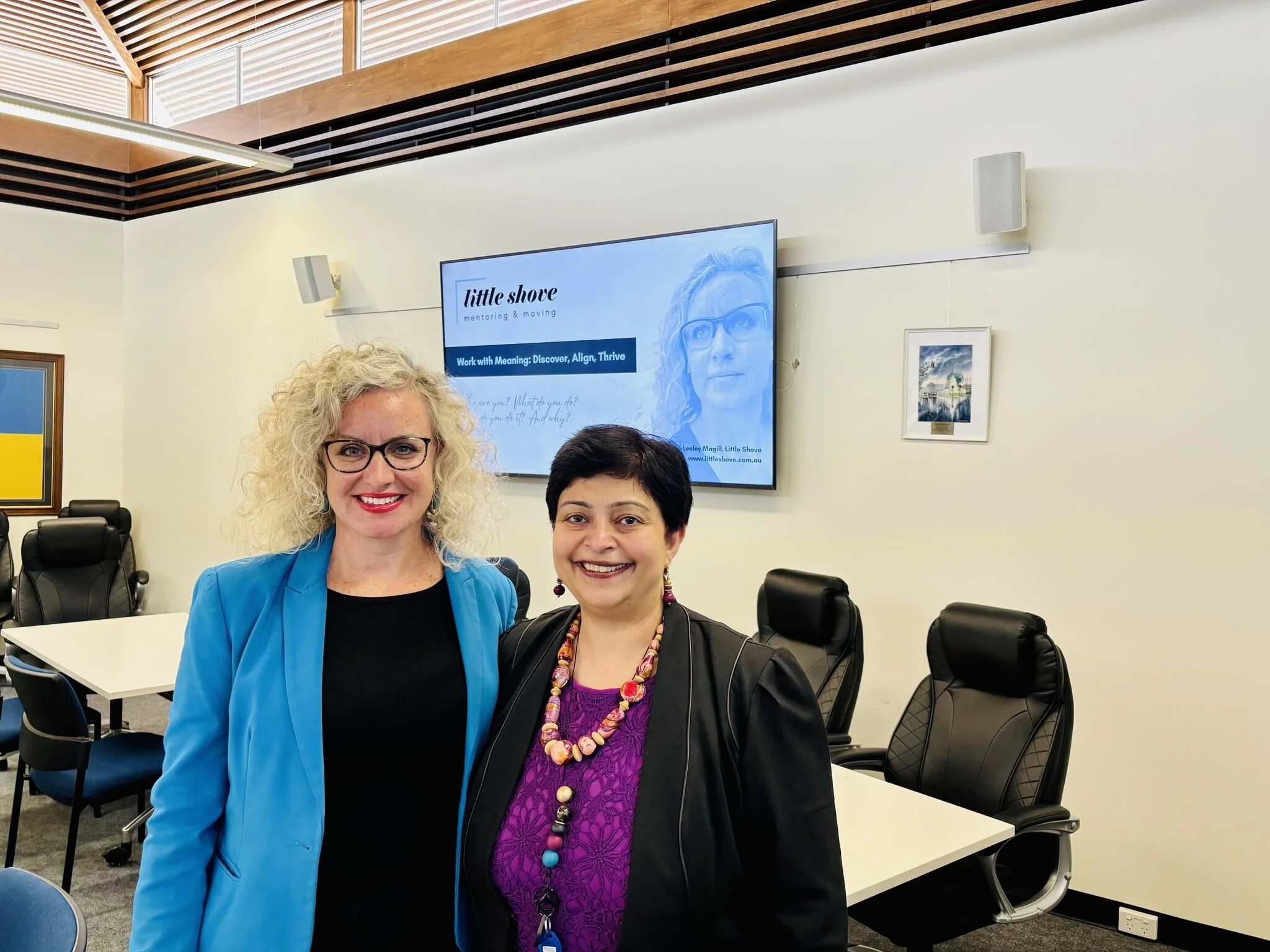 Two women smiling in a conference room, with a large screen behind them displaying a presentation titled 'little shove mentoring & moving'.