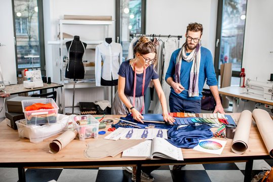 Two people working on fashion design projects in a bright studio, surrounded by fabric swatches, sketches, and sewing materials.