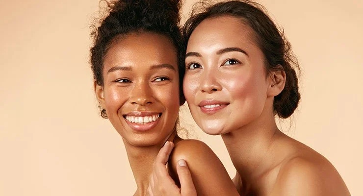 Two women smiling and close together, with one touching the other's shoulder, against a neutral background.