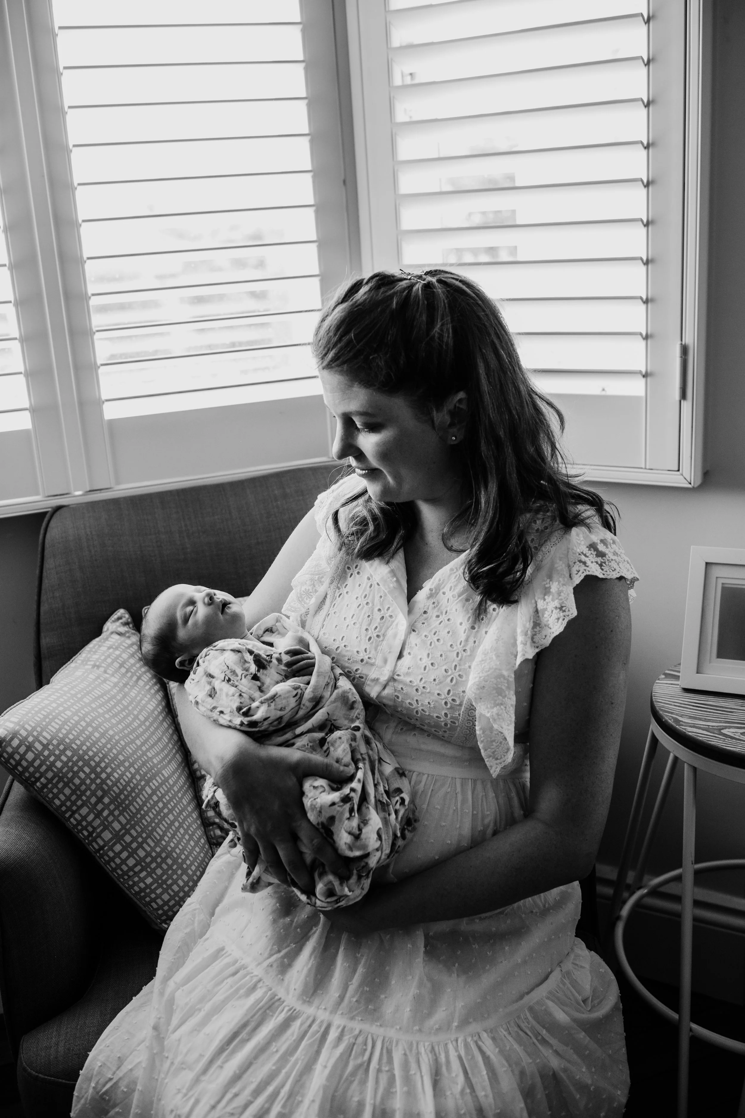 A woman sitting on a couch holding a newborn baby wrapped in a blanket, looking at the baby with a gentle expression, in a room with window shutters and a side table.