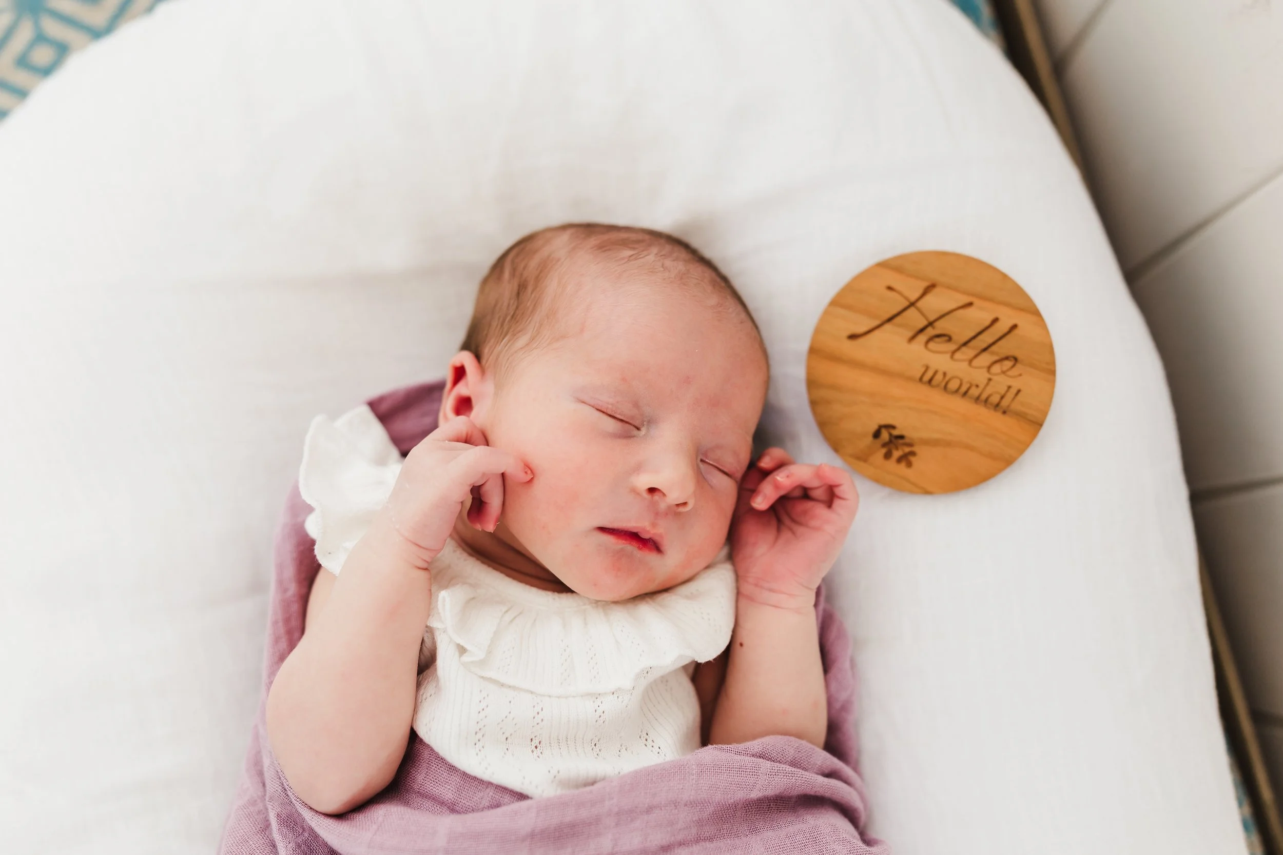 Newborn baby sleeping on a white surface with a round wooden sign that reads 'Hello world!'