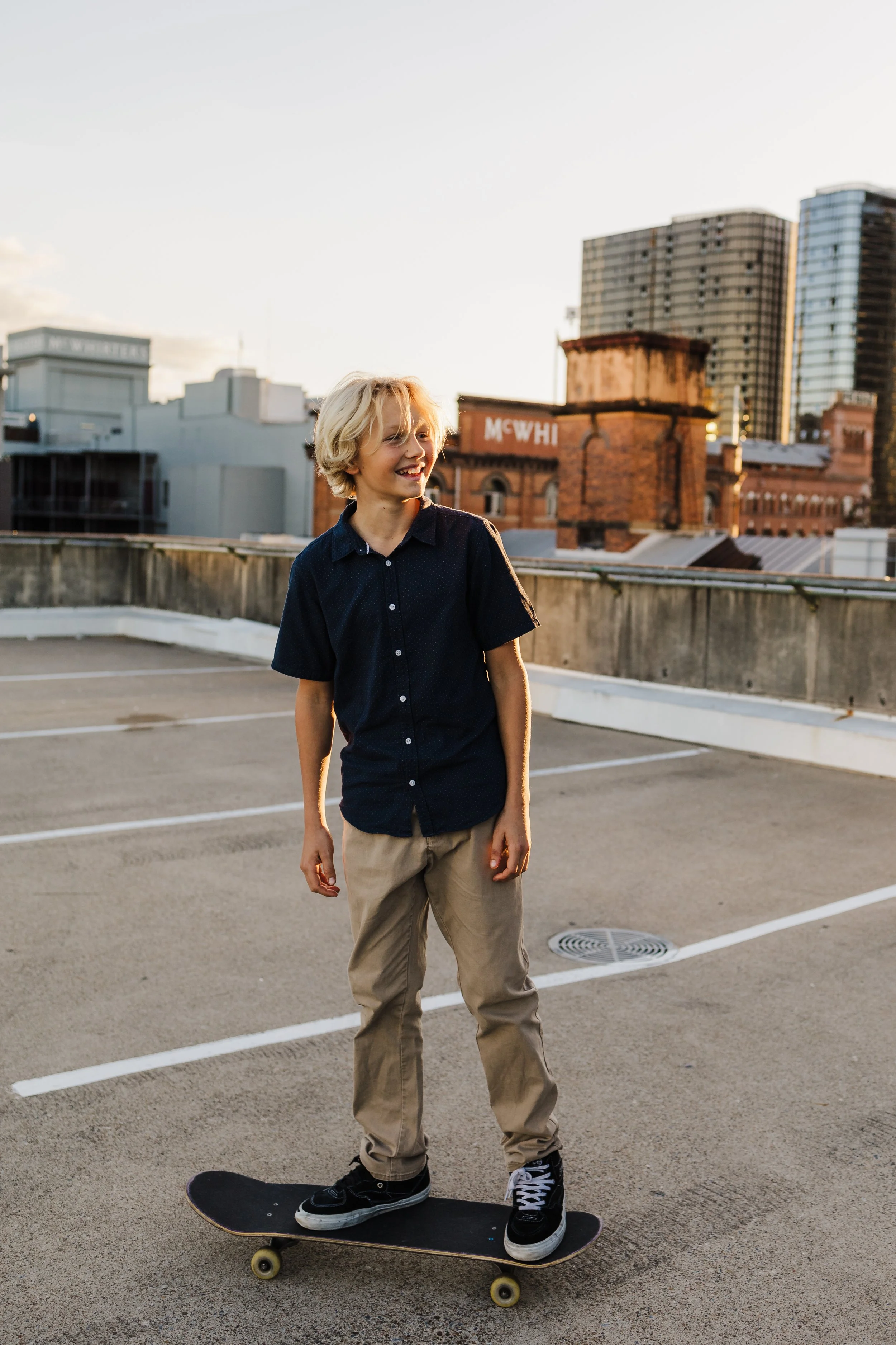 A young boy with blonde hair smiling while skateboarding on a rooftop parking lot during sunset, with city buildings in the background.