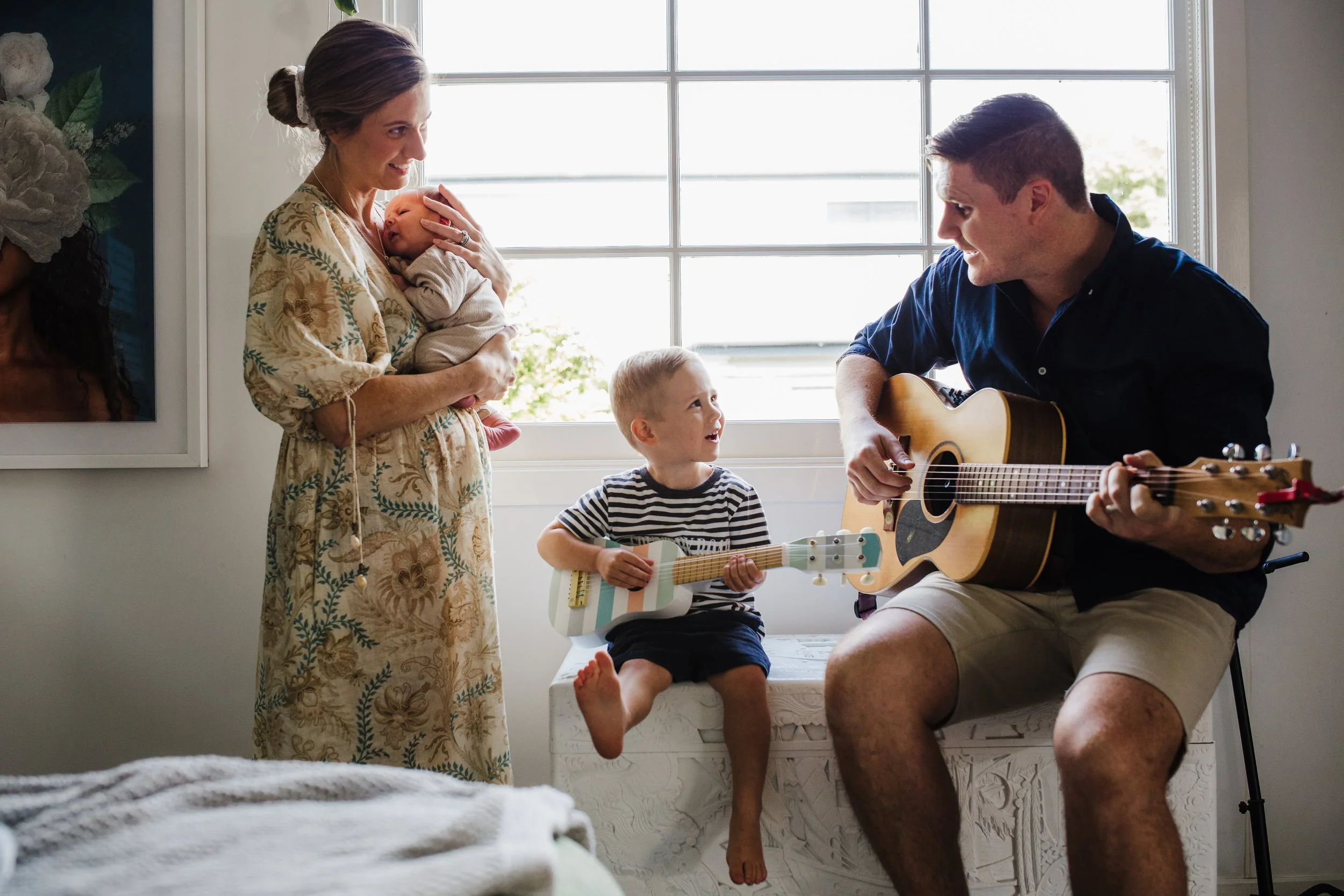 A family in a bright room, with a woman holding a sleeping baby, a young boy playing a colorful guitar, and a man playing an acoustic guitar, all engaging and smiling.