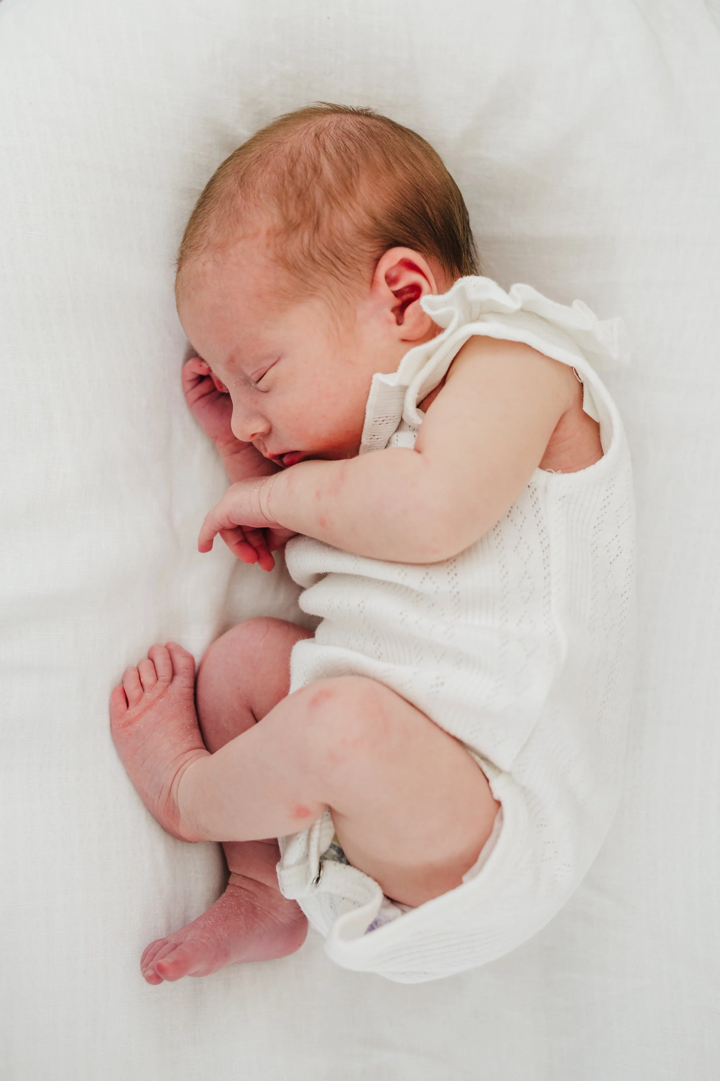 A sleeping newborn baby with light skin and reddish hair, lying on its side on a white blanket, wearing a white sleeveless top.