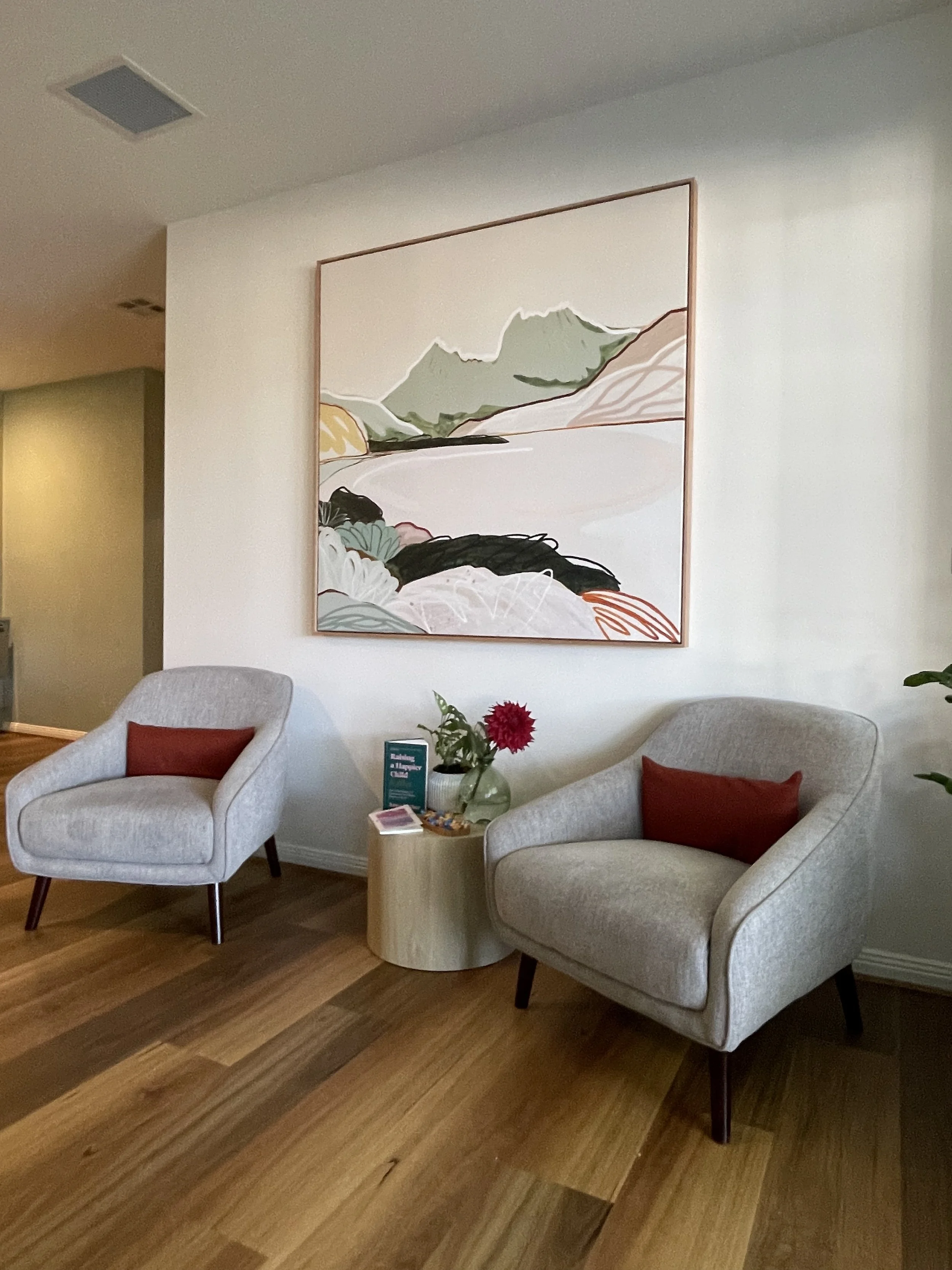 Waiting room with 2 grey chairs, side table with book and flowers, and artwork of a landscape behind