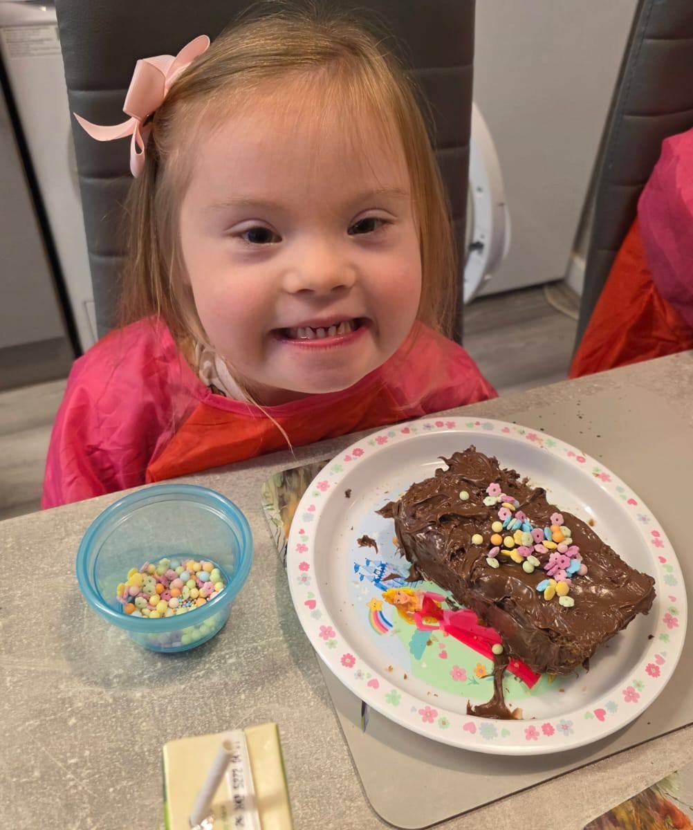 A young girl with Down's syndrome sits at a table, with a big piece of chocolate cake in front of her that she has decorated with chocolate icing and sprinkles. She is smiling and wearing a pink apron.