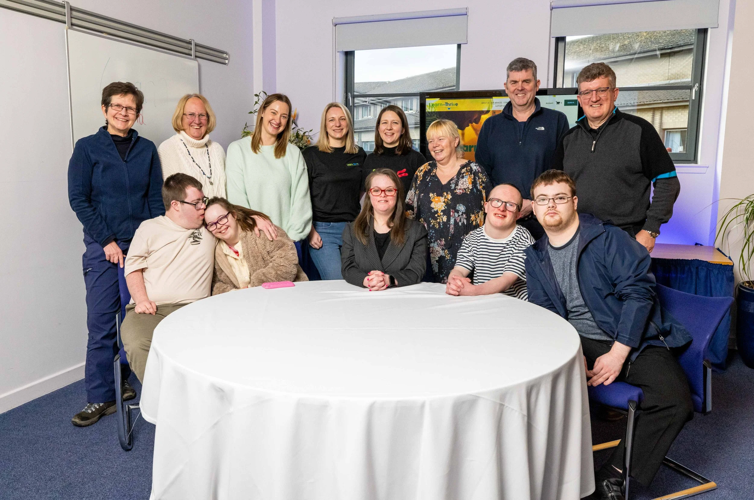 Five people with Down's syndrome sit around a table. Parents and staff stand behind them. Everyone is smiling.