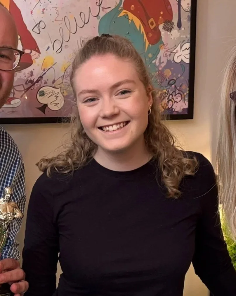 Smiling young woman with curly blonde hair wearing a black long-sleeve shirt, standing indoors with colorful artwork on the wall behind her