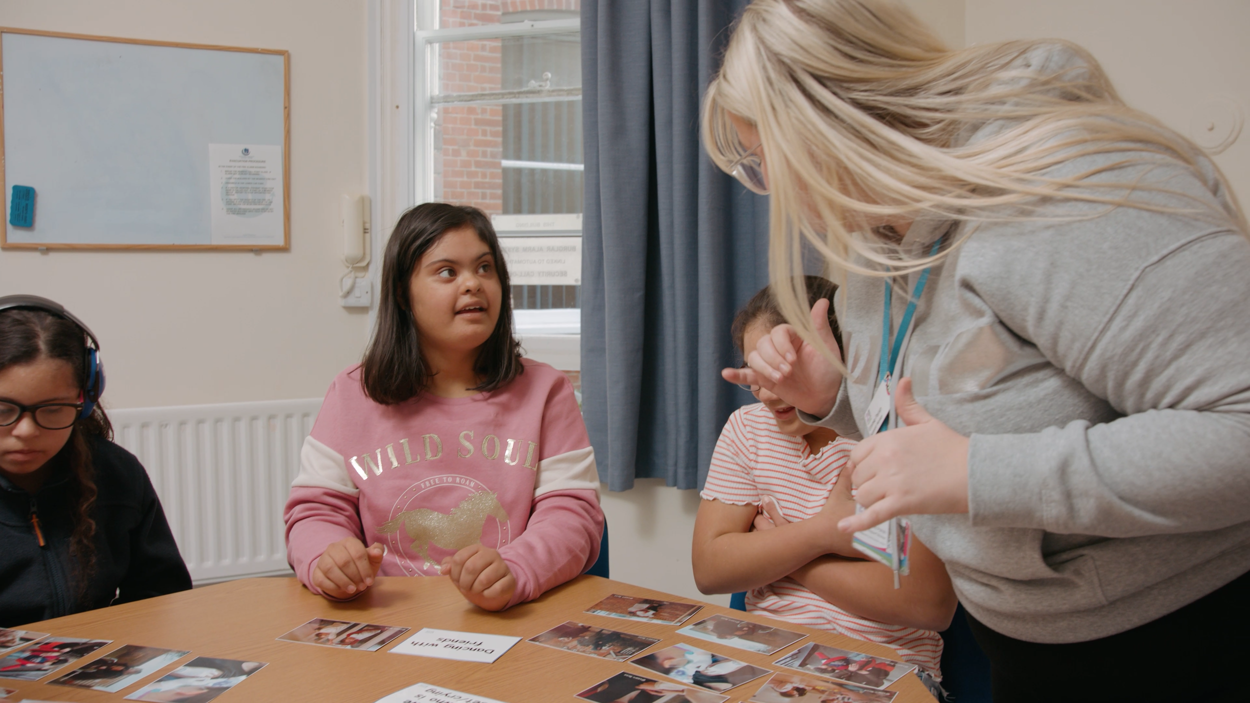 Three girls with Down's syndrome are being taught by a specialist teacher at a 21 Together session. The girl on the left wears headphones, the girl in the middle is looking and listening to the teacher, and the teacher is in front of the other girl.