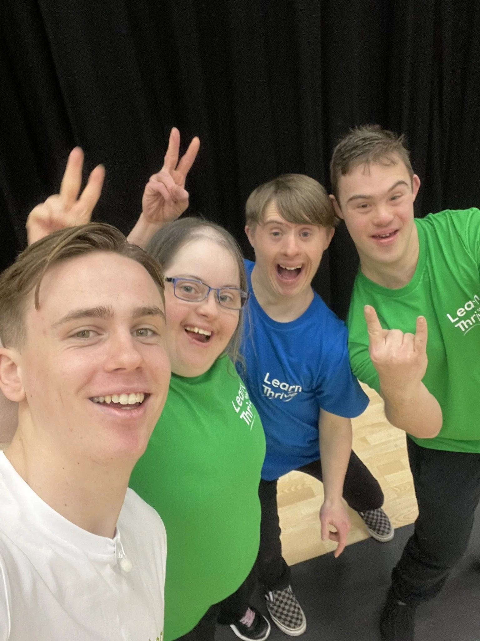 Group of four young people taking a selfie together, smiling and making gestures with their hands, indoors against a black background.