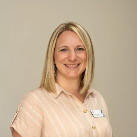 A smiling woman with blonde hair wearing a beige button-up shirt against a plain background.