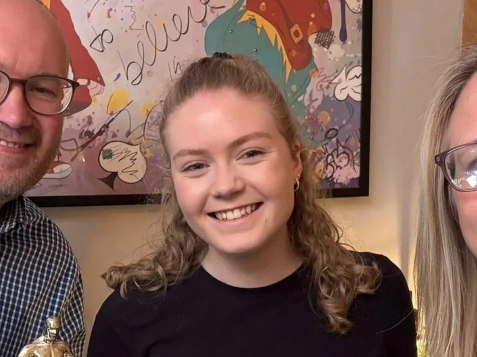 A smiling young woman with curly hair and earrings is standing next to a man and a woman, in front of colorful, abstract artwork.