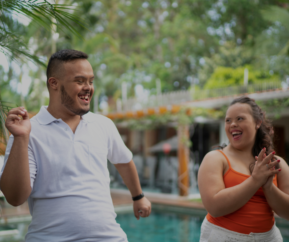 a man and a woman with down's syndrome are dancing and smiling at each other. they are in a tropical location.