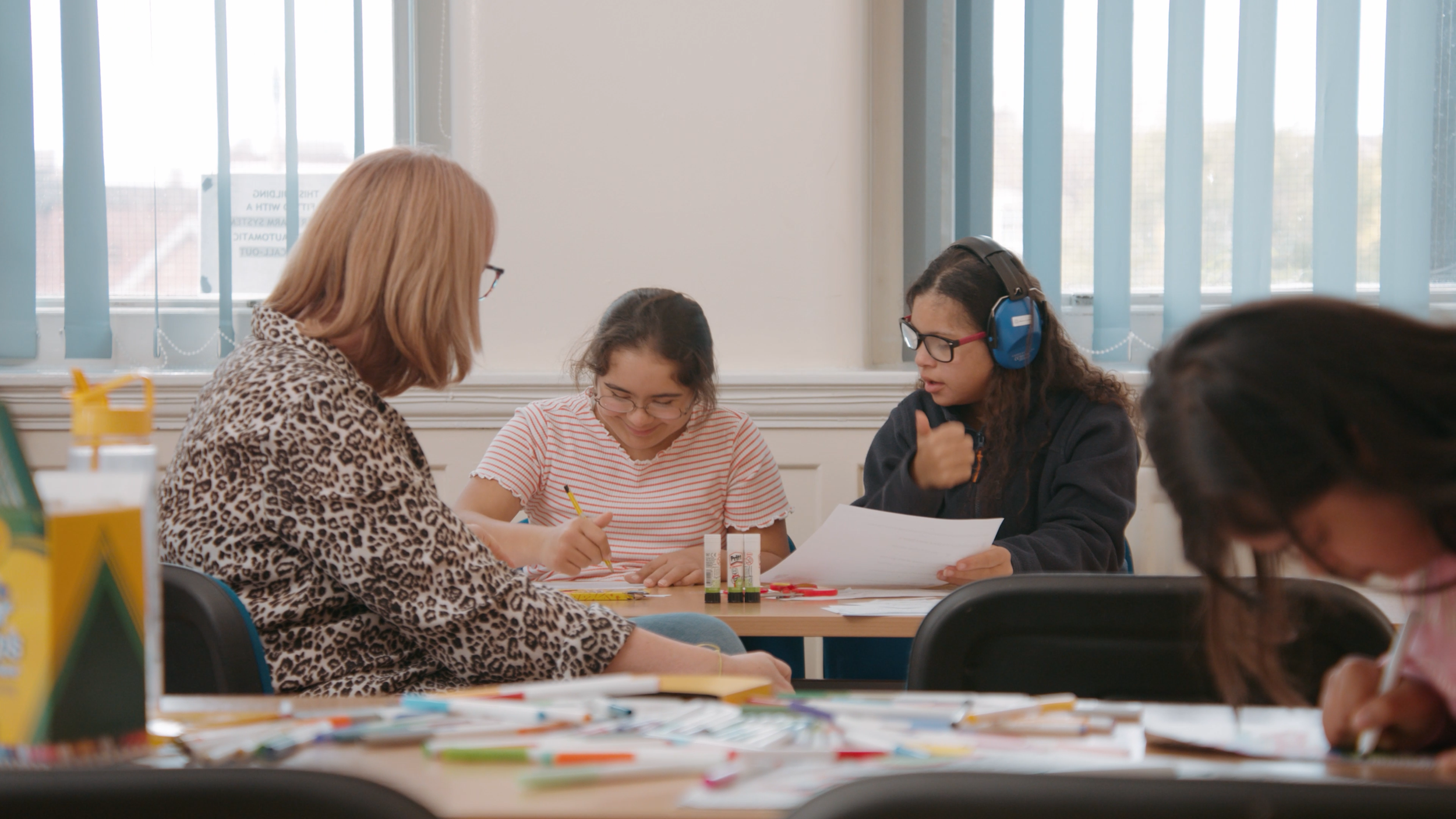 Two girls sit at a desk working through worksheets. Their teacher sits in front of them.