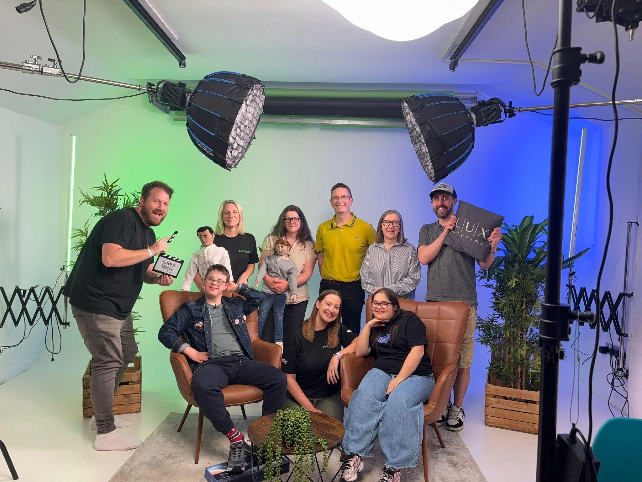 A group of people smile at the camera, sitting on a film set. There is a green blue light behind them.