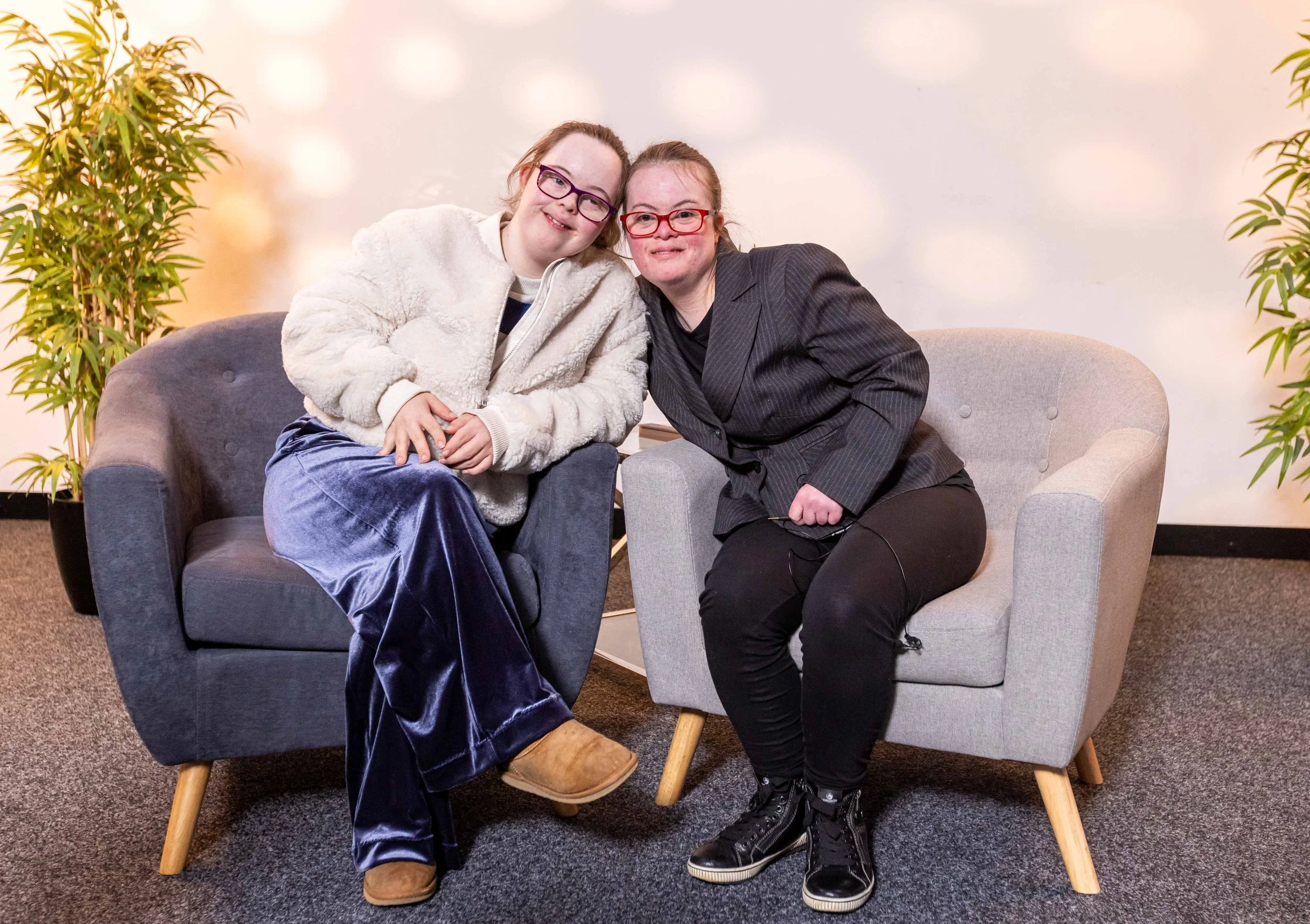 Two young women sitting on a gray armchair, smiling. One wears glasses, a cream-colored furry jacket, and blue velvet pants. The other wears glasses and a dark pinstripe blazer. They are indoors, with potted plants on each side and a neutral wall background.