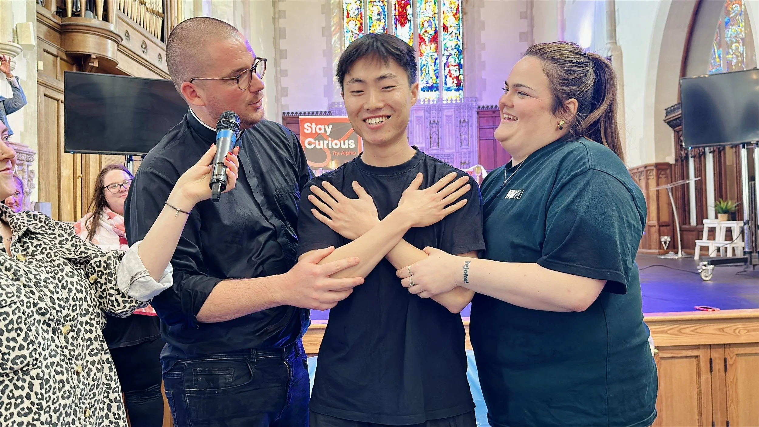 A group of people in a church, including a smiling young man with cross-arms and a woman holding his arm, being interviewed with a microphone. Others are watching in the background.