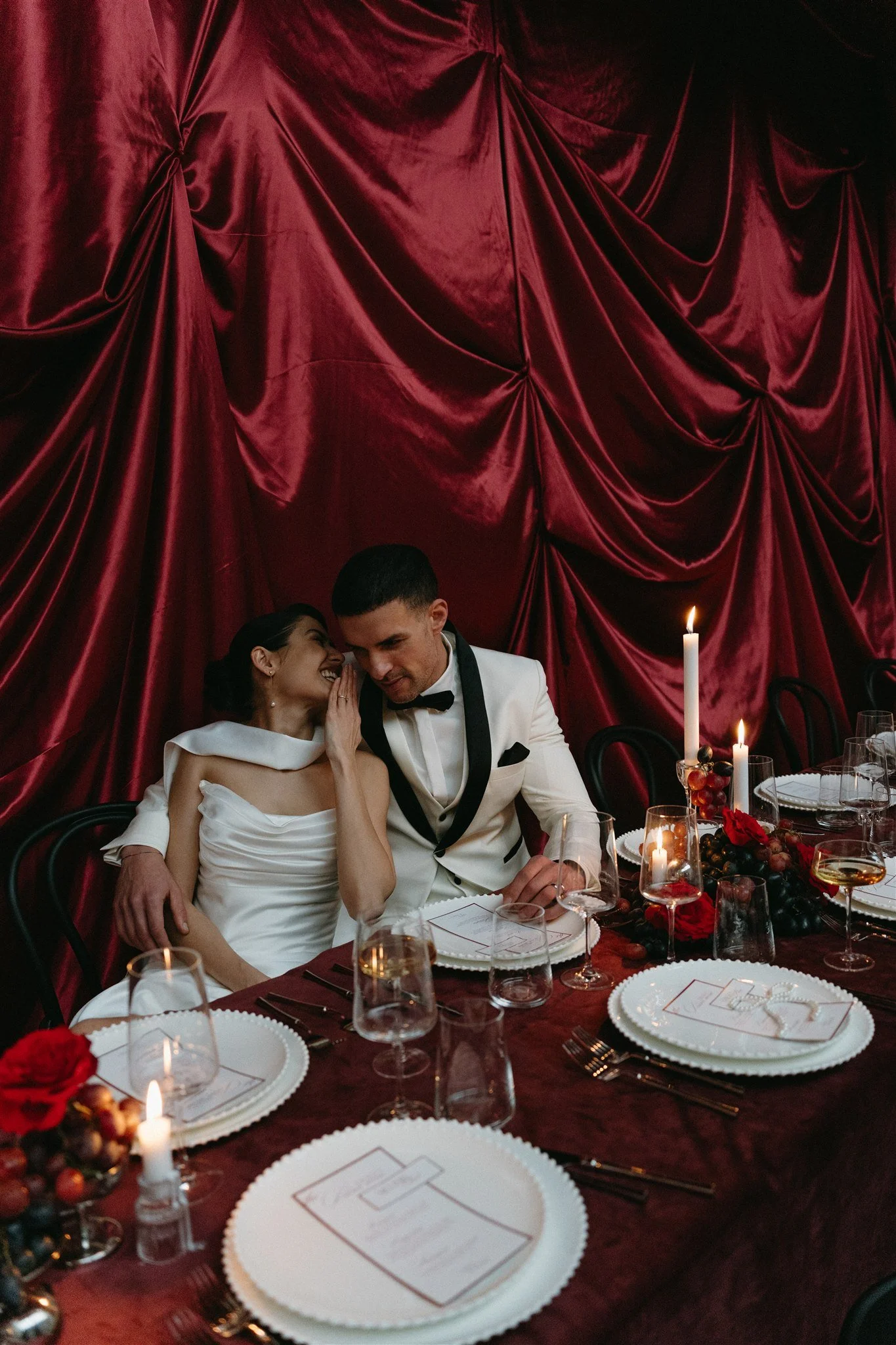 Opera masquerade wedding table setting. A couple of bride and groom sitting on the table. The bride is laughing and whispering something to the groom. The Background is full of red wedding ceiling draping, photographed by Joana Buchmayer a destinatio