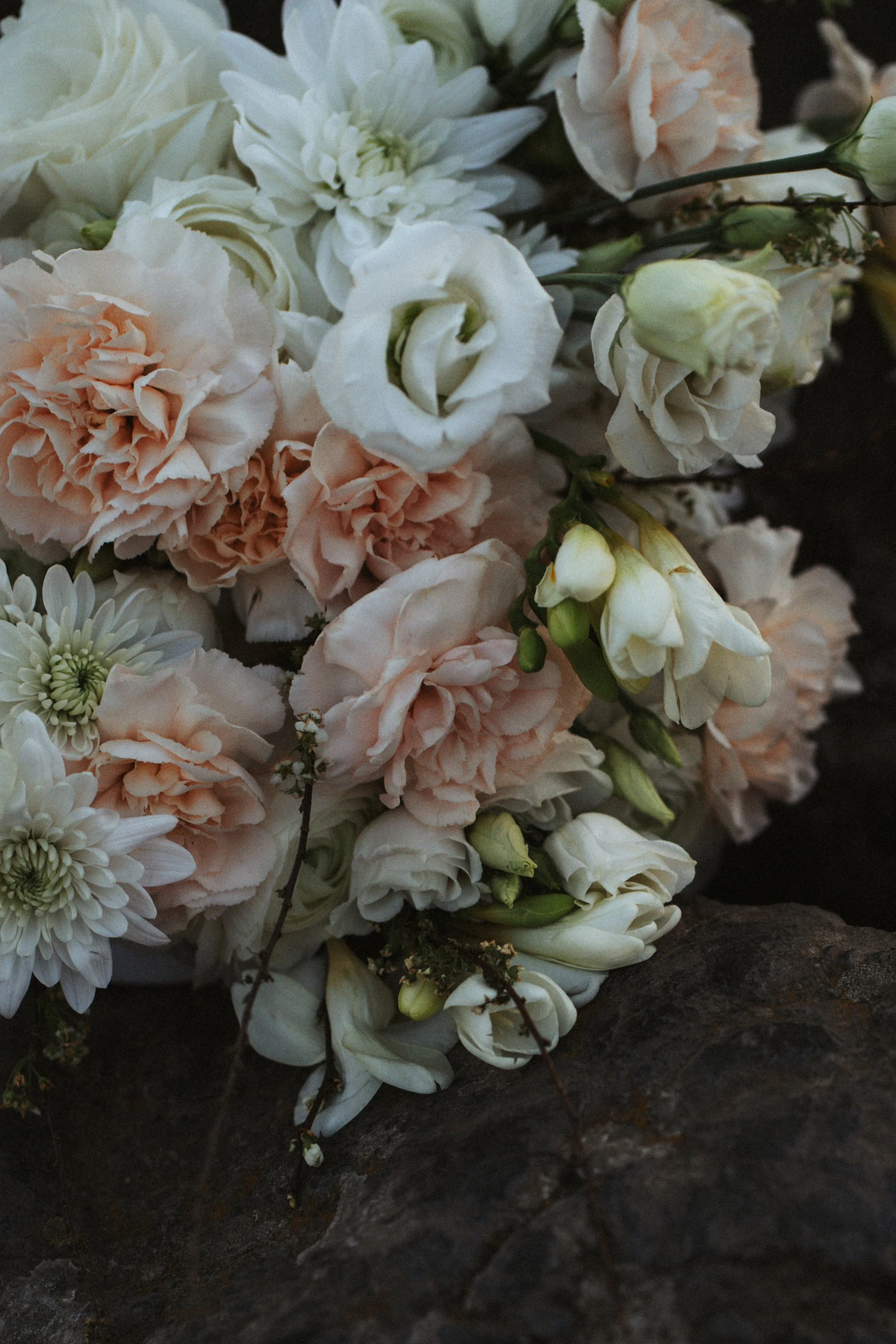 An exquisite arrangement of soft peach and white flowers nestled against a dark rocky backdrop, emphasizing nature's raw beauty alongside elegant styling, captured by Joana Buch, a distinguished artistic wedding photographer