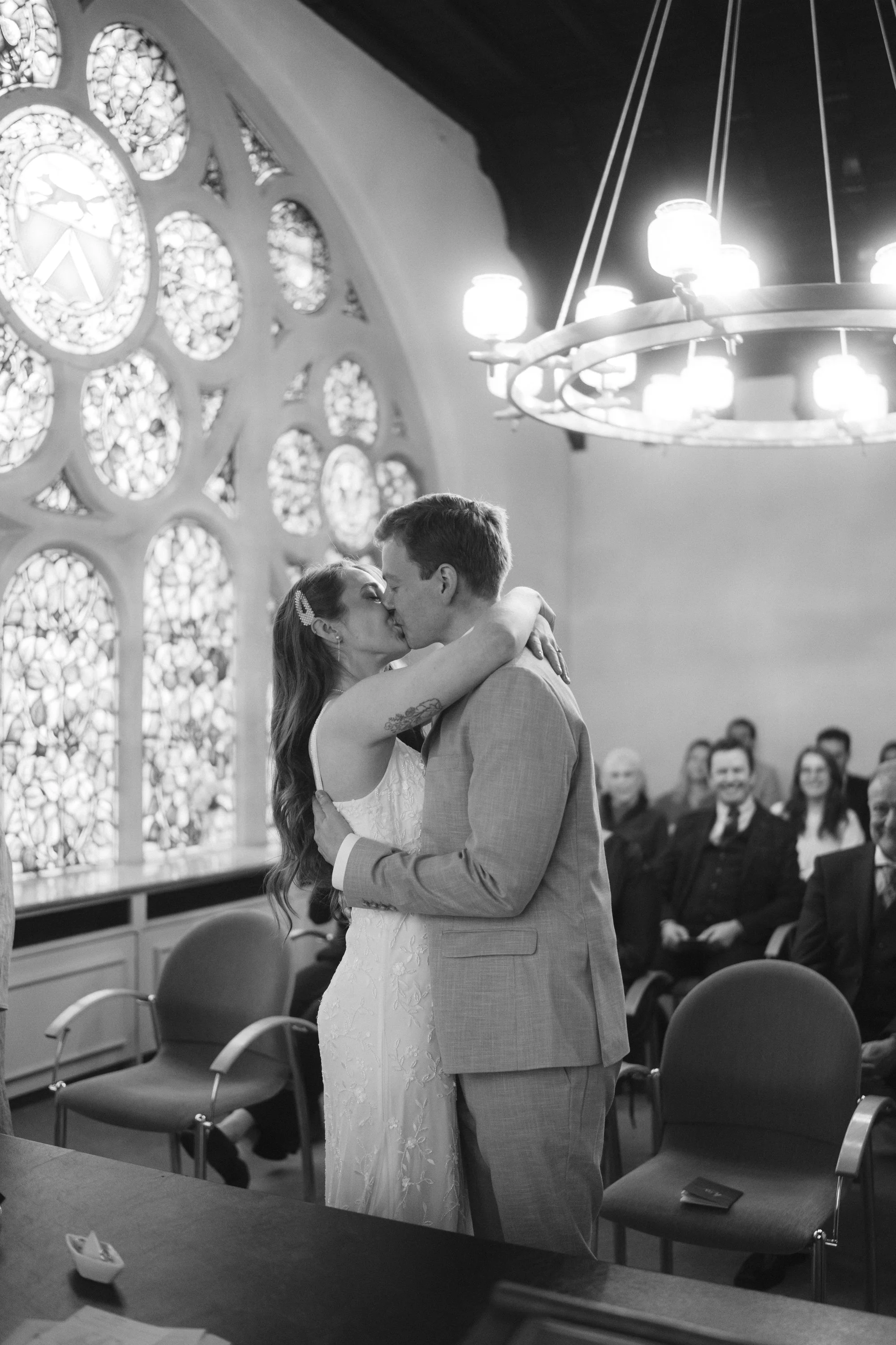 Standesamtliche Trauung in Wuppertal Vohwinkel, Bild zeigt die Aufnahme eines Paares welches sich nach dem JA Wort den ersten Kuss gibt, im Hintergrund sieht man die lachende Familie und eine wunderschönes Buntglasfenster