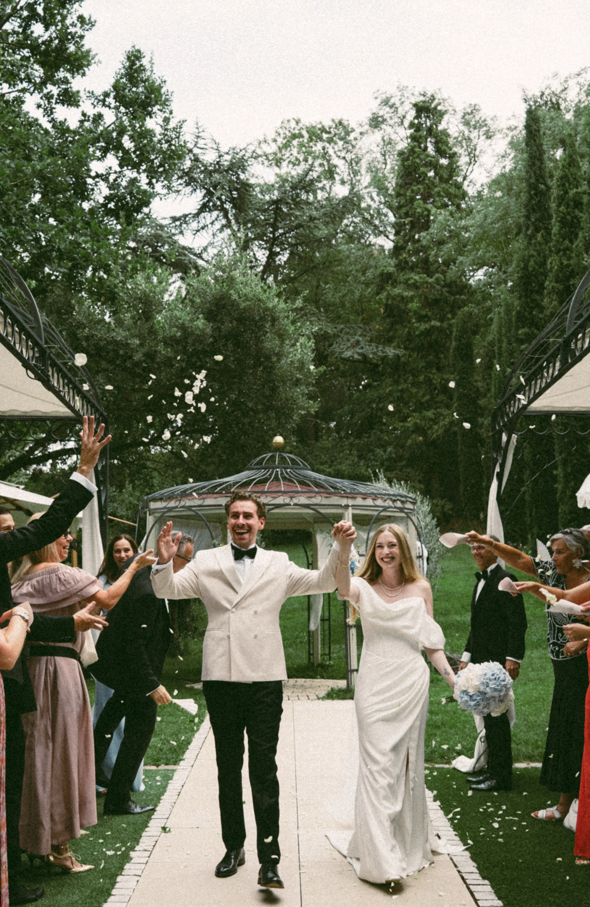 Pictures show a couple in beautiful black tie wedding outfit, the bride is wearing a satin slip dress. they getting celebrated from family and friend who throughing flowers over their head, when thy leaving the wedding ceremony.