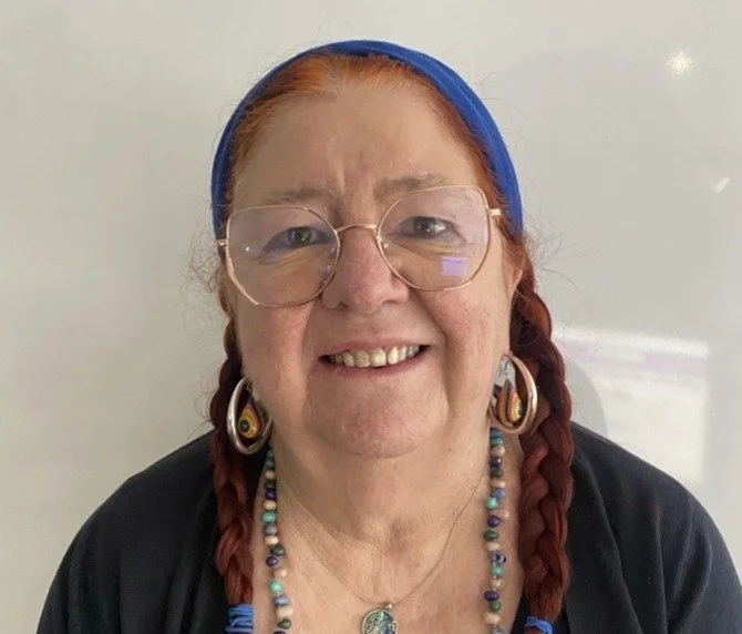A woman with red hair, wearing a pink headband, purple scarf, and black leather jacket, smiling outdoors near a body of water.