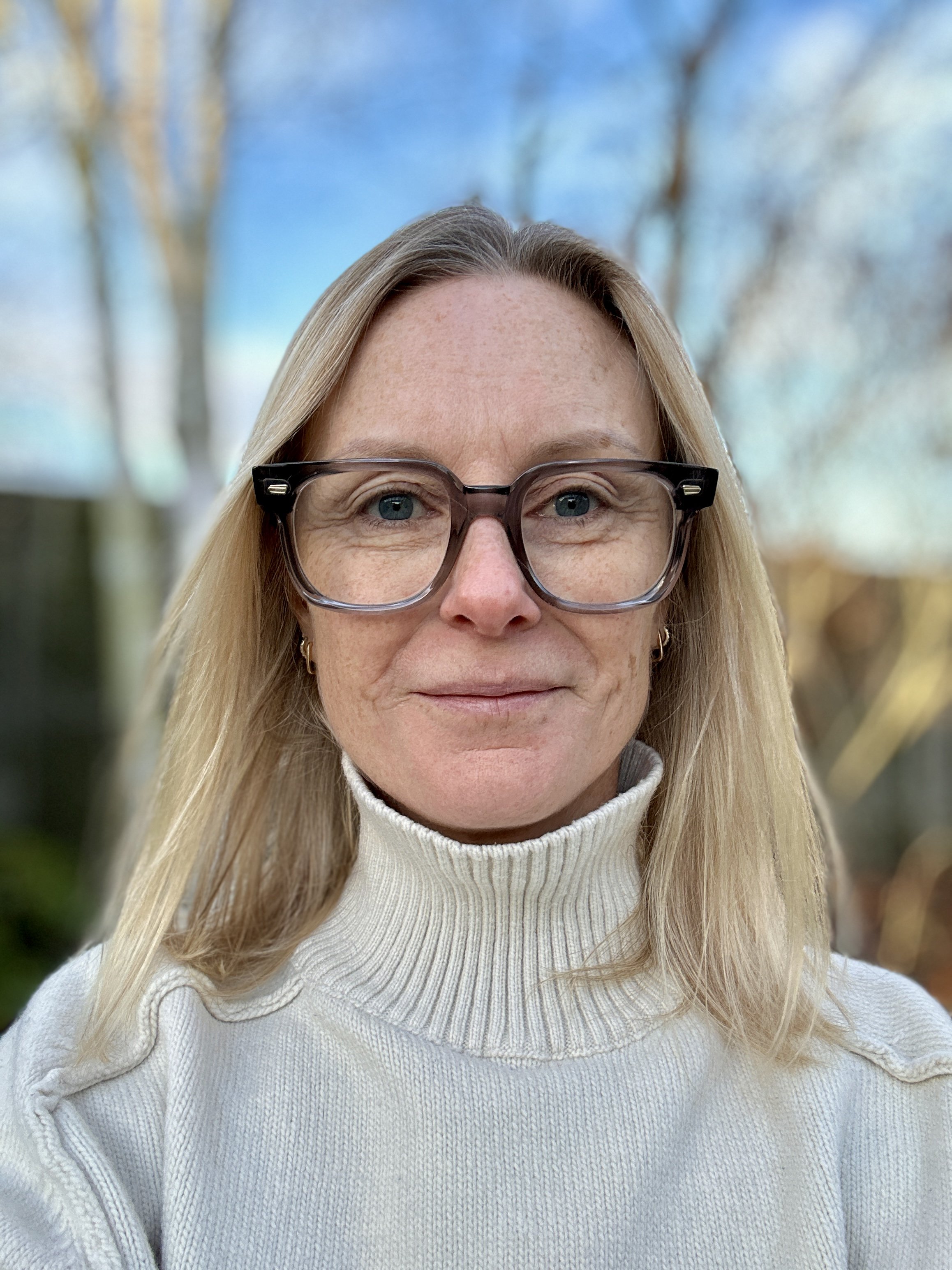 A smiling man with dark hair wearing a black sweater and a collared shirt, standing against a beige wall.