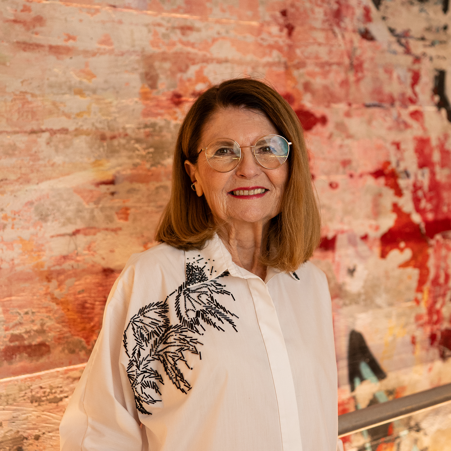 A woman with shoulder-length hair, wearing glasses, a white blouse, and a skirt, seated in front of a dark background with concentric white circles.