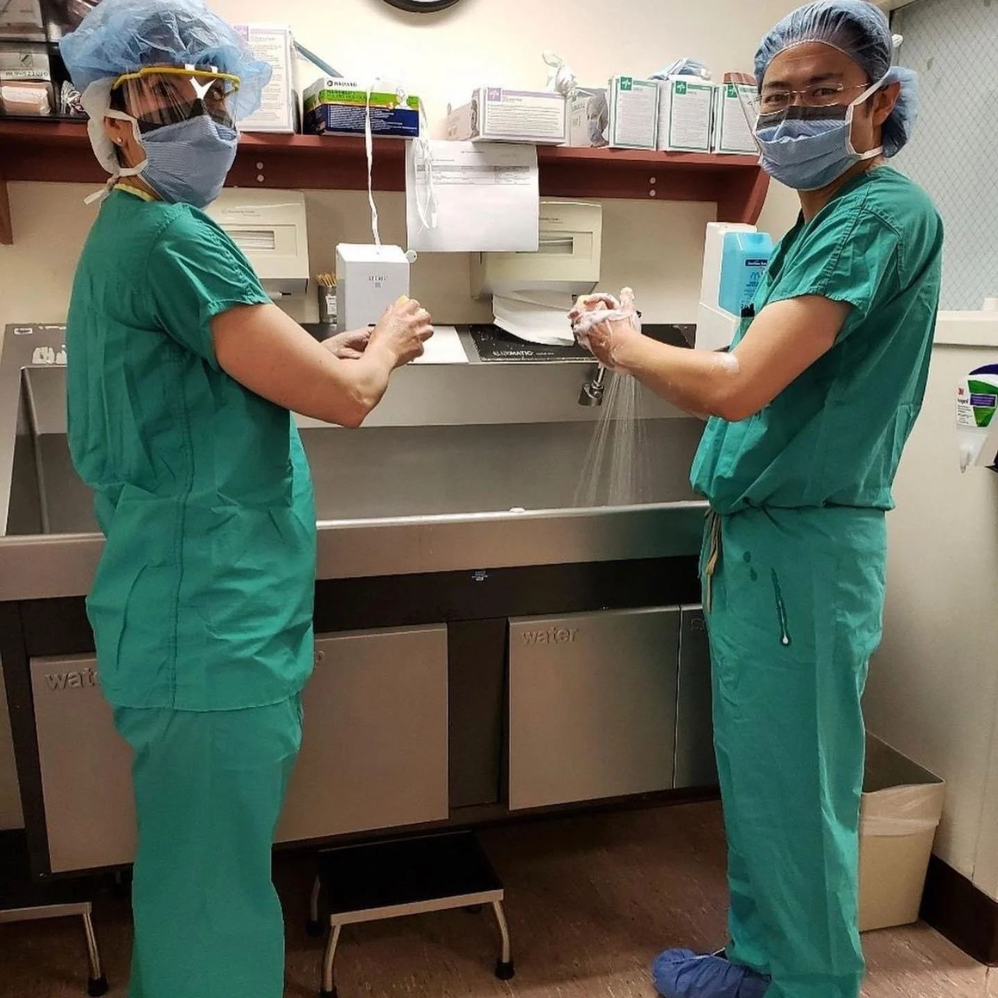 Two healthcare professionals in scrubs, masks, and protective gear washing hands in a hospital or clinical setting.