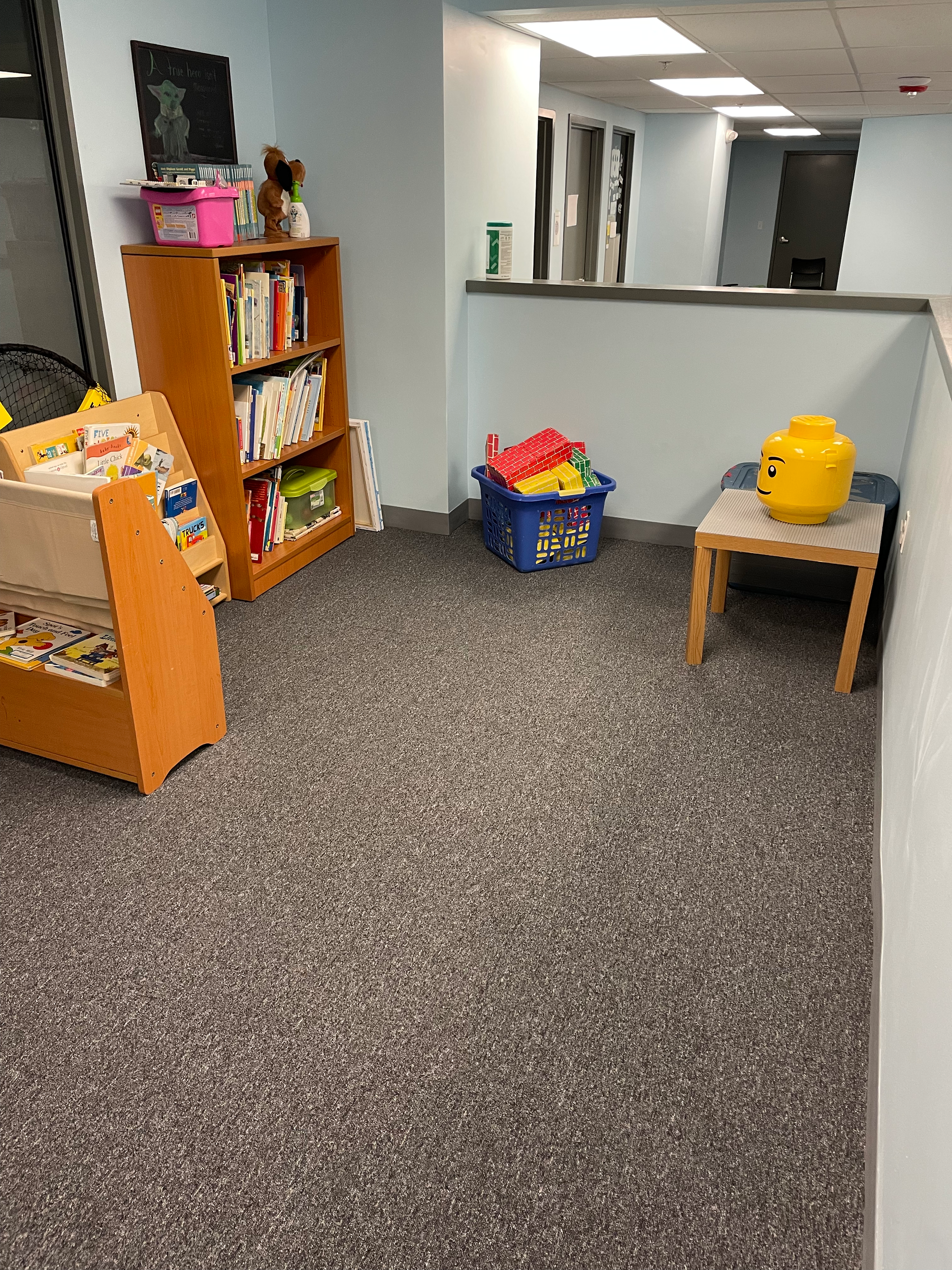 an open space with bookshelves, a table and a basket of toys.  There is a yellow lego head storage box on the table.  The floor has grey carpet and the walls are light blue.