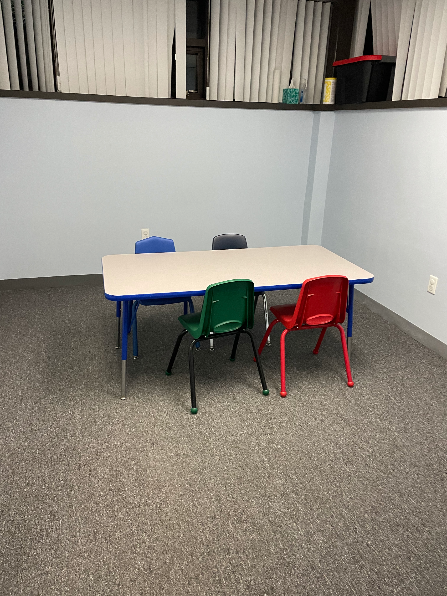 A room with a kids table and four chairs - one red, one blue, one green and one black.  There is a high corner window with shades.  The carpet is grey and the walls are light blue.
