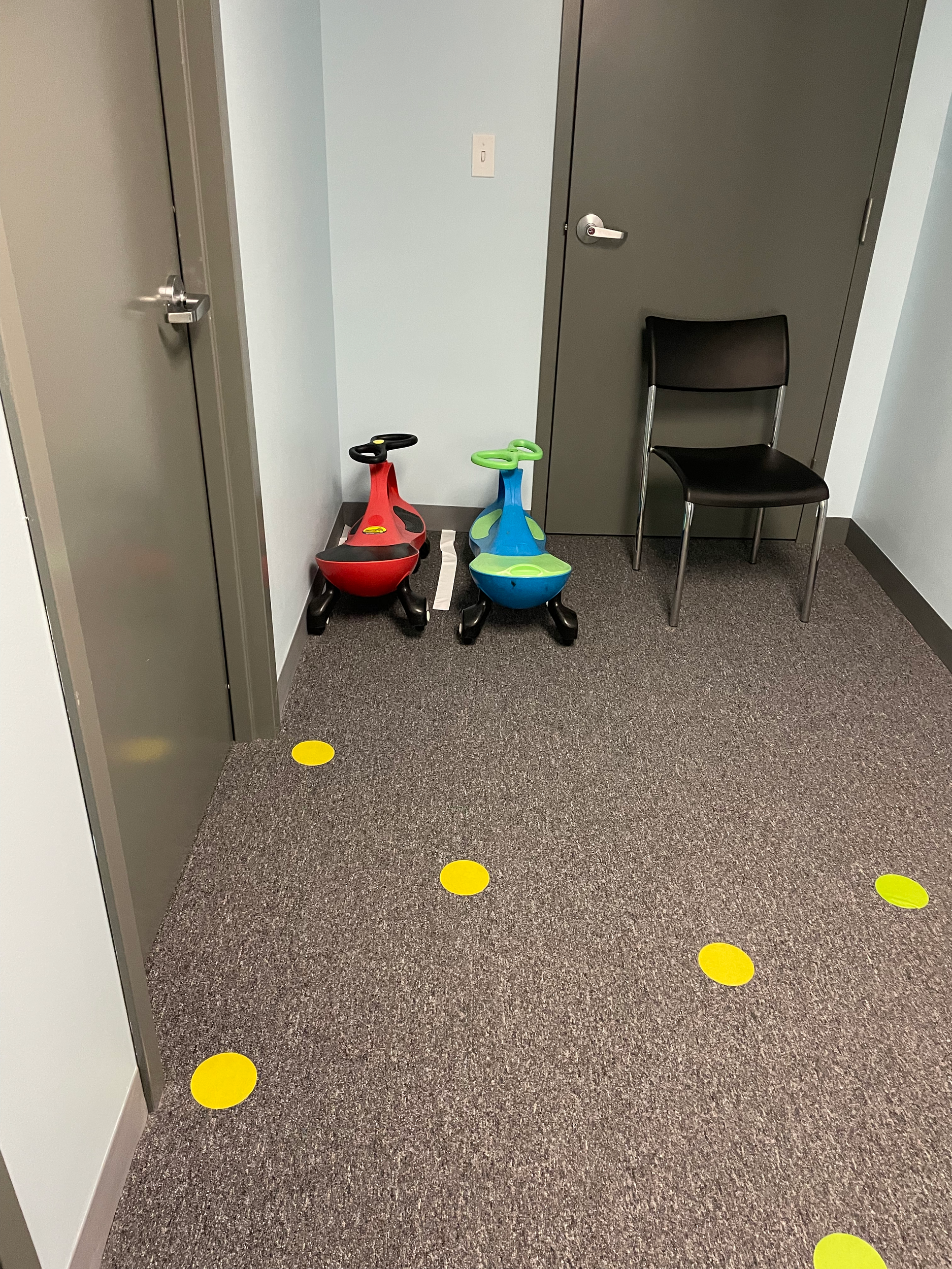 a hallway with two grey doors and grey carpet and blue walls. There is a black chair in the back right corner and two kids sit-on toys.  There is also large yellow dots on the floor.