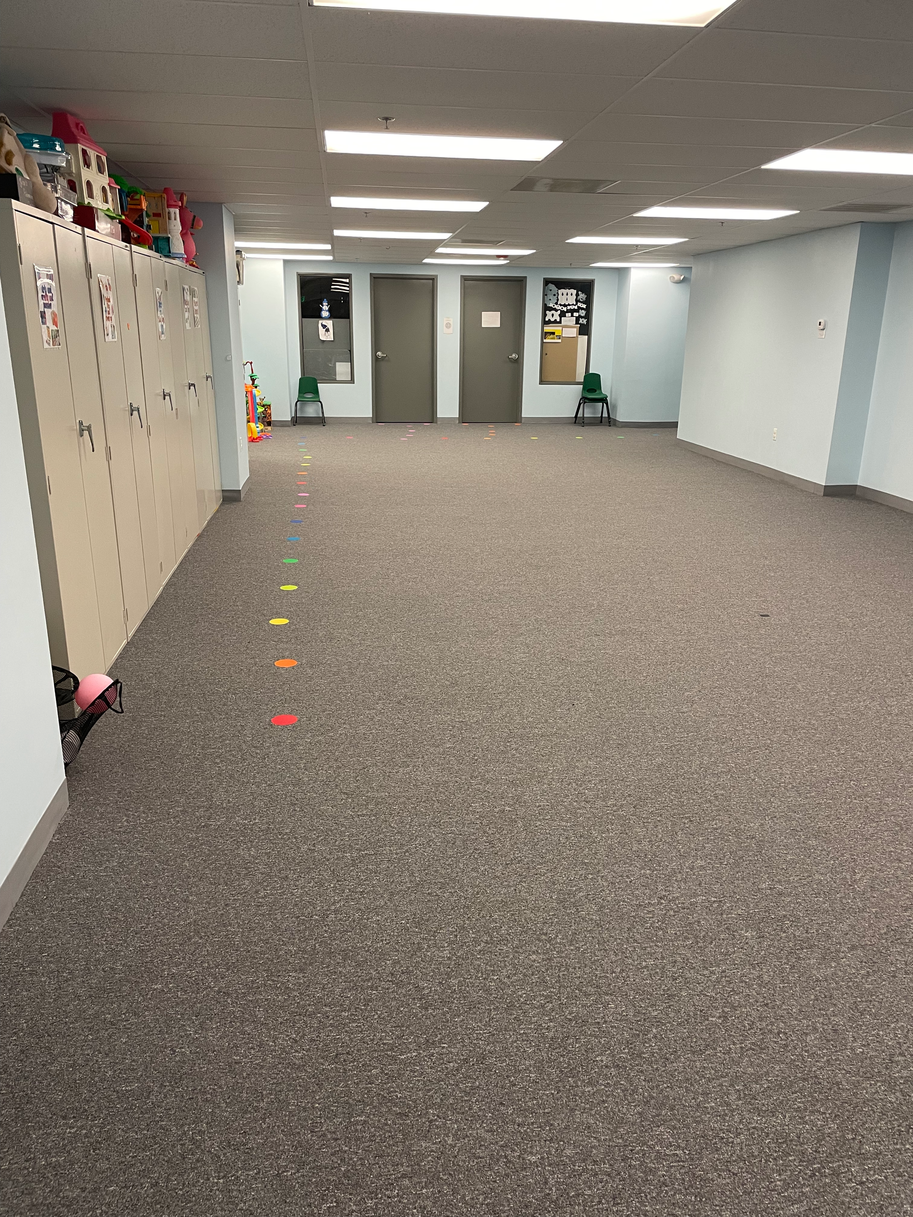 A large room with grey carpets and blue walls.  There are storage cabinets all along the left side of this open space.  To the back there are two rooms with grey doors.  There are some large dots in different colors on the floor alongside the cabinet