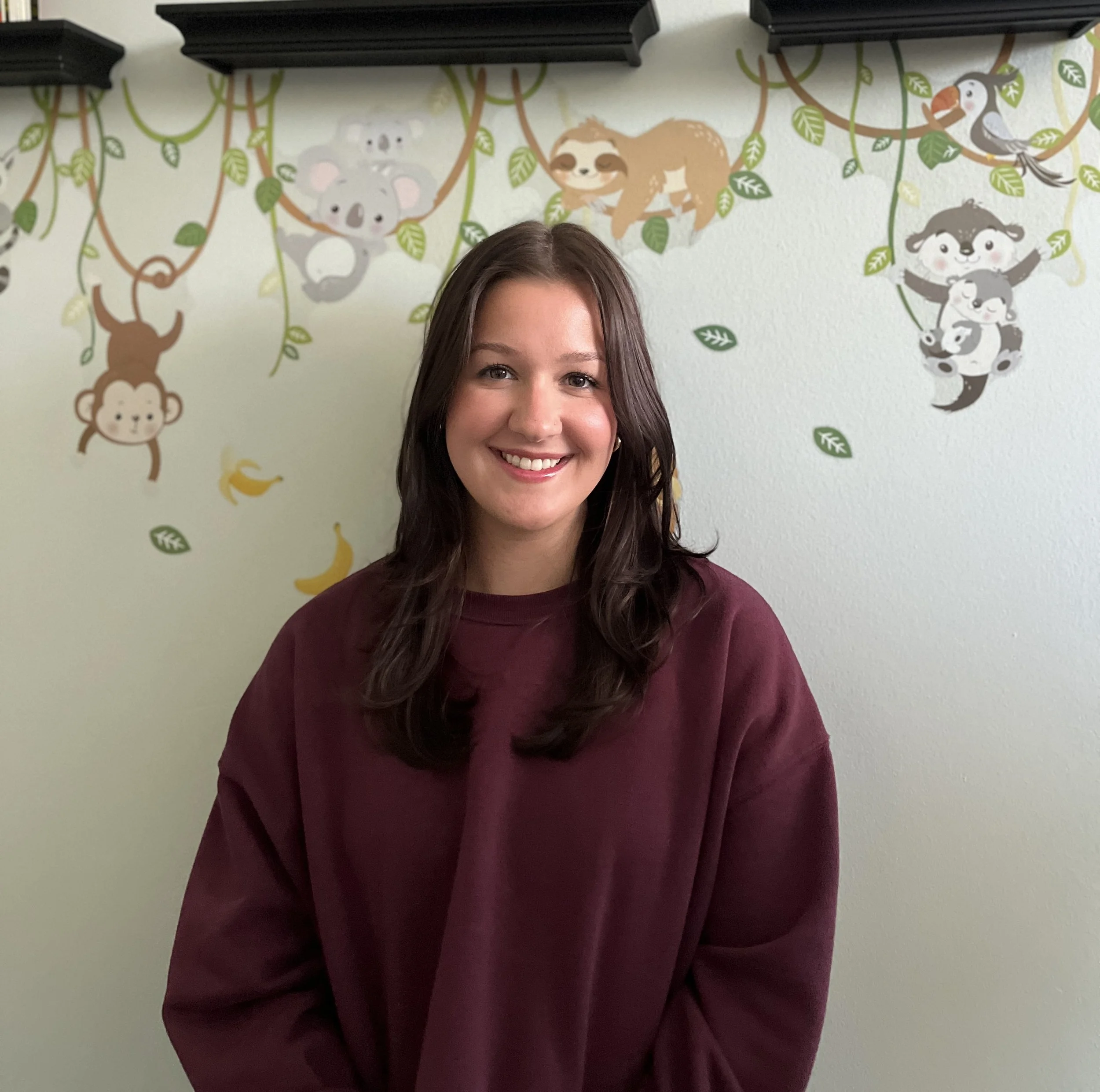 Amanda is smiling and wearing a burgundy top and is standing against a wall with some zoo animals hanging from vines.