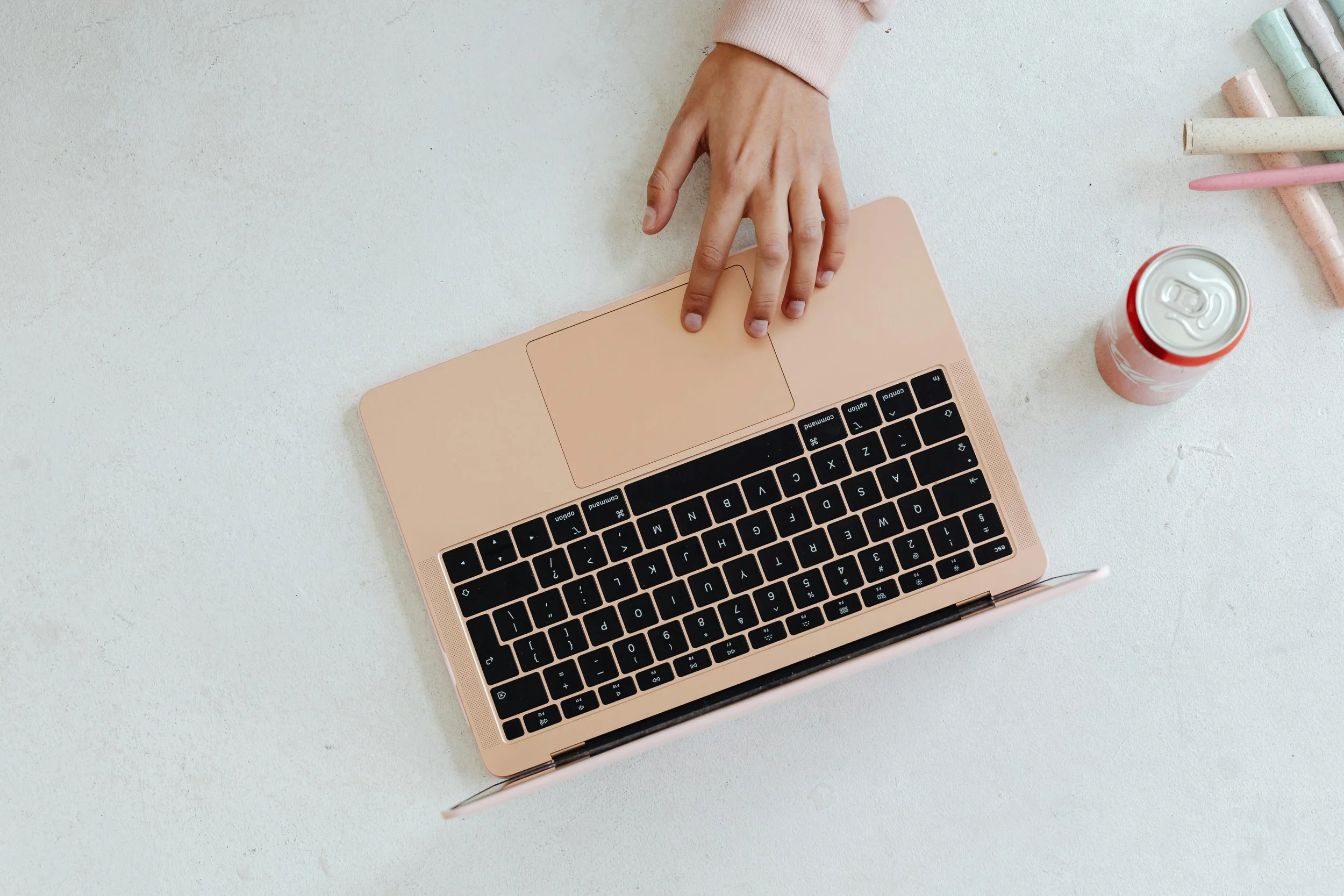 Top view of a person's hand using a pink laptop on a white surface, with a pink beverage can and pastel-colored chalks nearby. Virtual trauma therapy to Ohio and Kentucky.