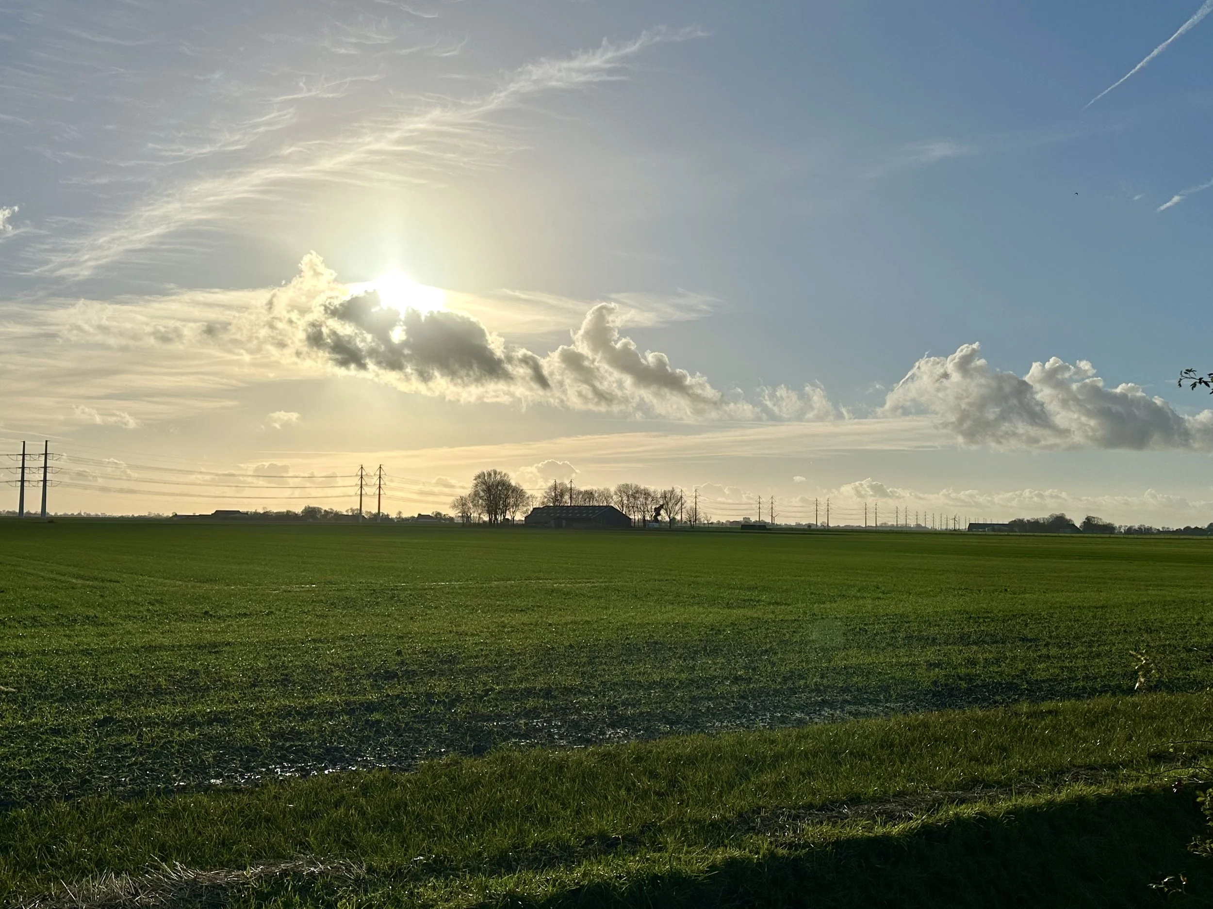 Een uitgestrekt groen veld onder een heldere blauwe lucht met wolken en de zon die door de wolken schijnt.
