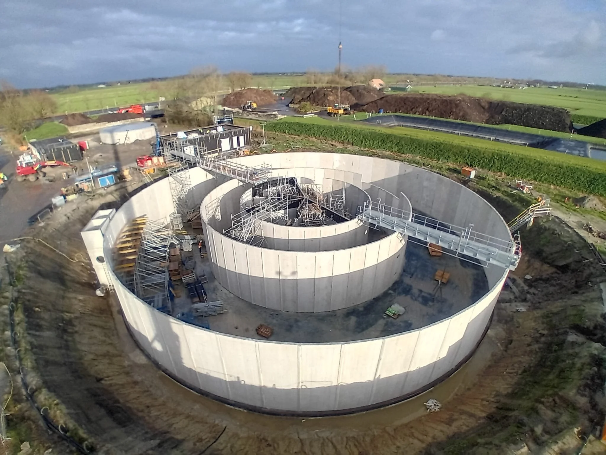 Een bouwplaats waar een grote, ronde cementen structuur wordt gebouwd, omringd door steigers en bouwmaterialen, met een groen landschap en een bewolkte hemel op de achtergrond.