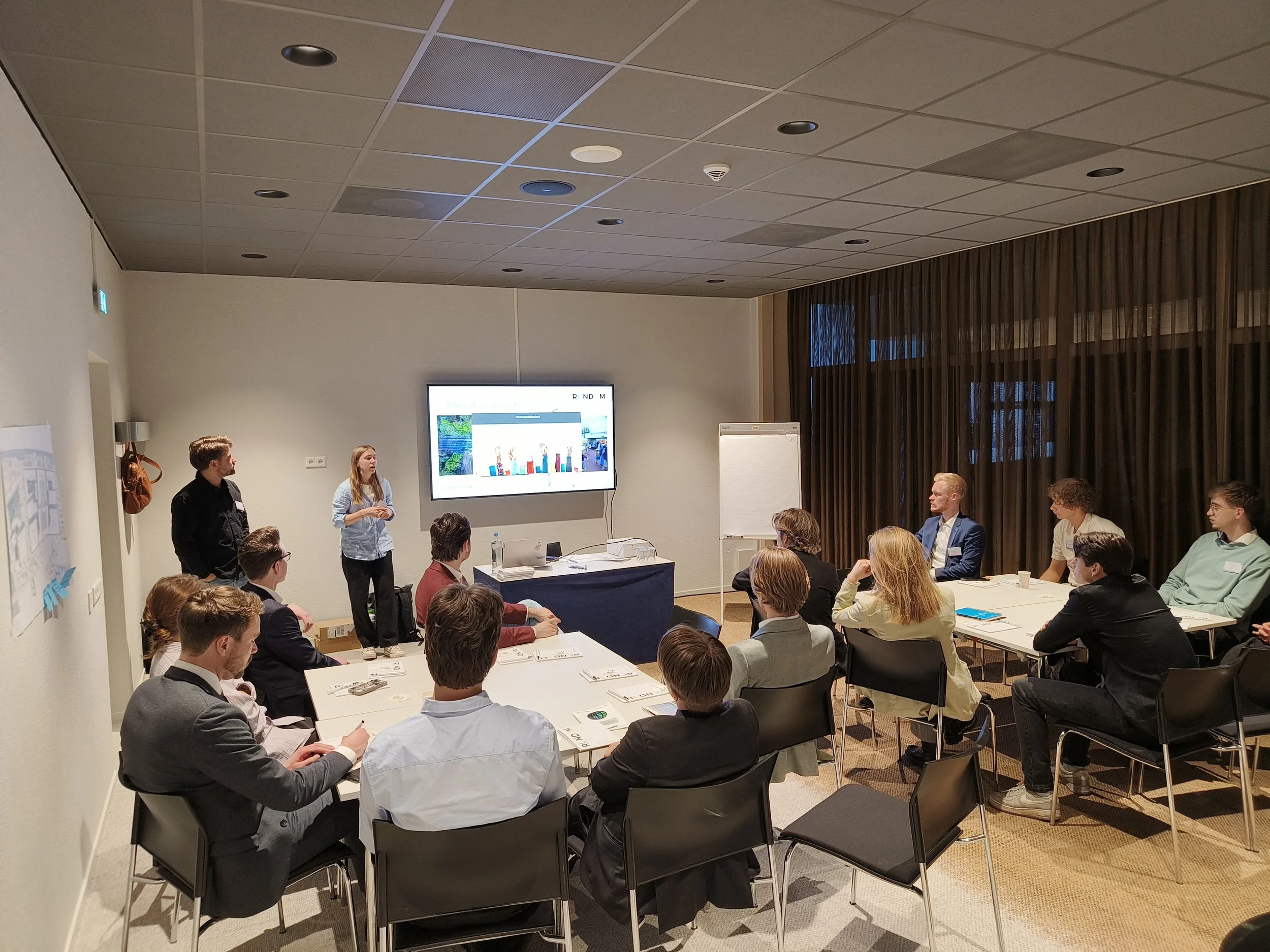 Een groep mensen zit aan tafel tijdens een presentatie in een vergaderruimte, met een vrouw die spreekt voor een scherm en een man die naast haar staat.