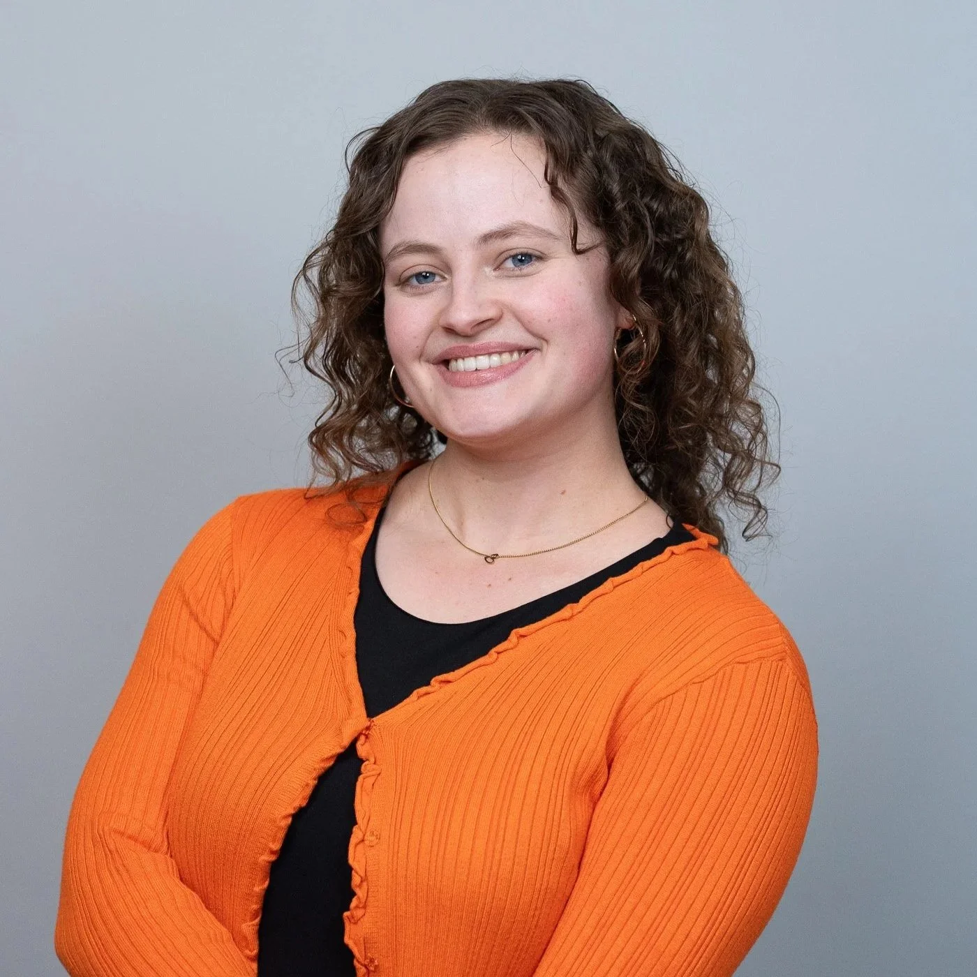 Portret van een lachende vrouw met curly haar, zwart T-shirt en oranje cardigan, staand tegen een lichtgrijze achtergrond.