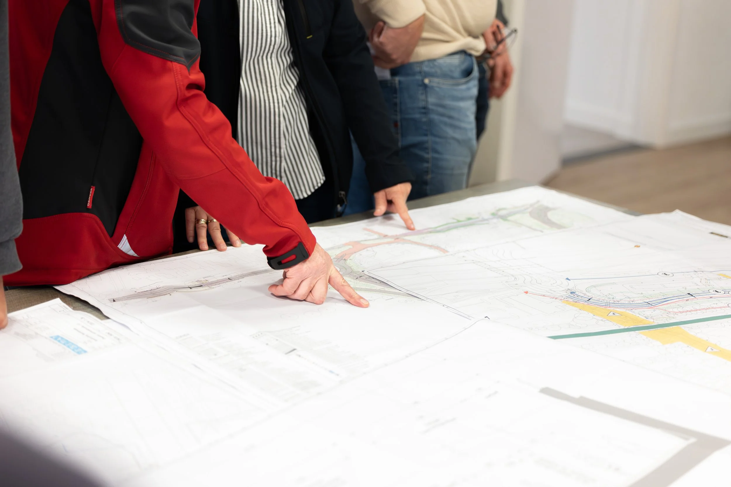 Mensen bekijken gerenommeerde technische tekeningen en plattegronden op een grote tafel tijdens een bespreking.