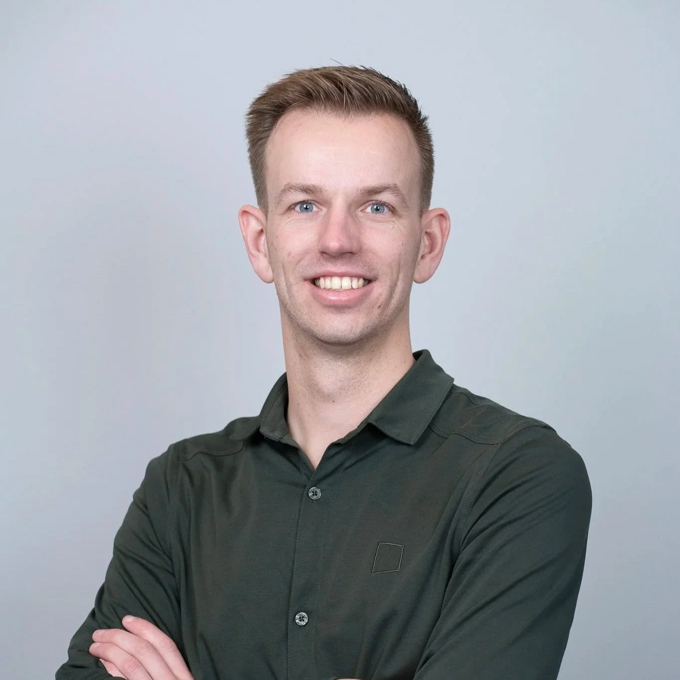 Portretfoto van een jonge man met kort, lichtbruin haar en een brede glimlach, gekleed in een donkergroene blouse, staande voor een lichtgrijze achtergrond.