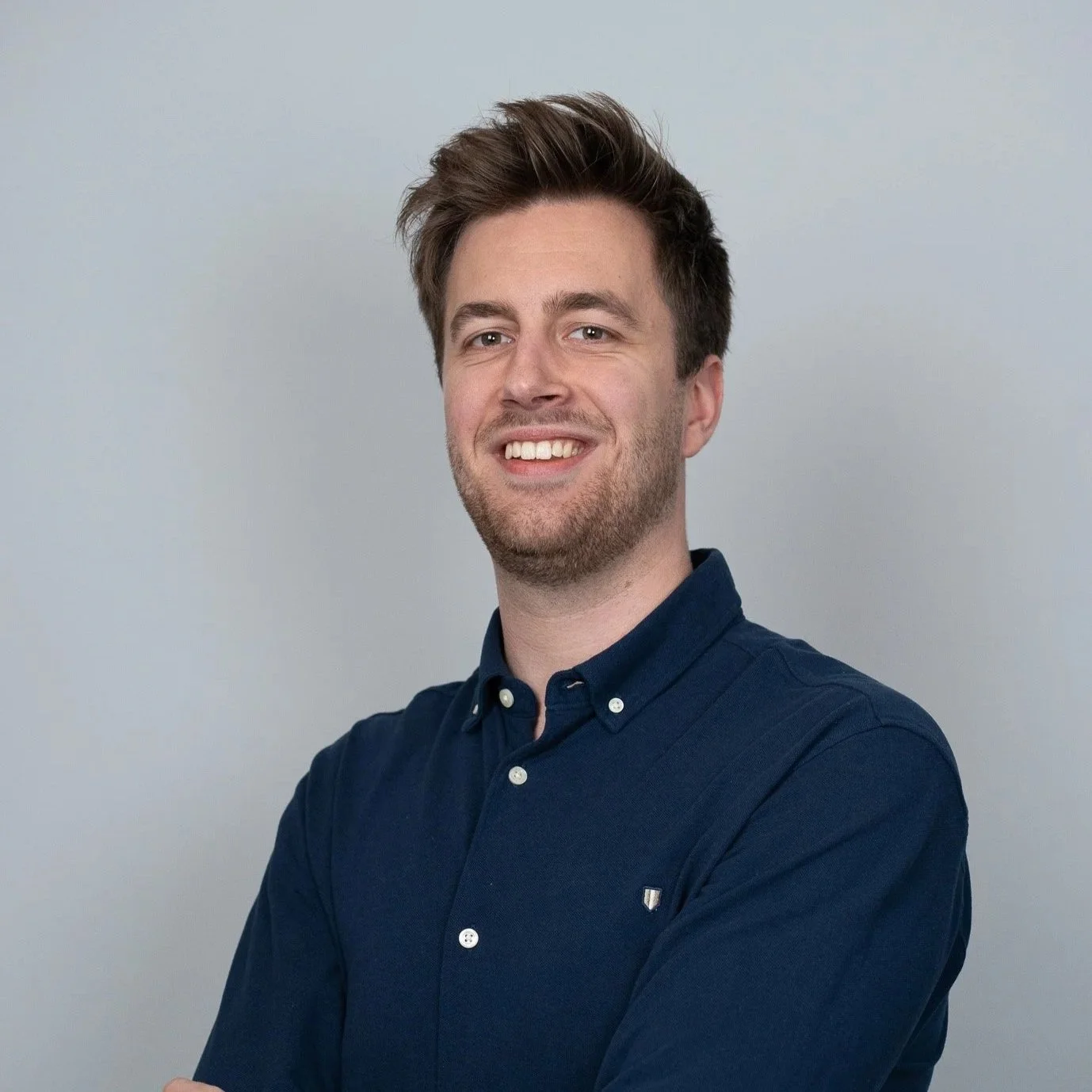 Portret van een jonge man met bruin haar, gekleed in een donkerblauw overhemd, lachend tegen een lichte neutrale achtergrond.
