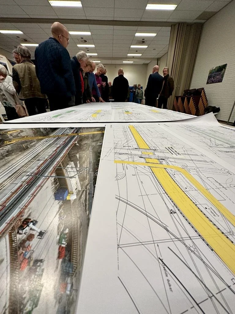 Groep mensen die naar grote technische tekeningen of bouwplannen kijken op een tafel in een verlichte zaal.