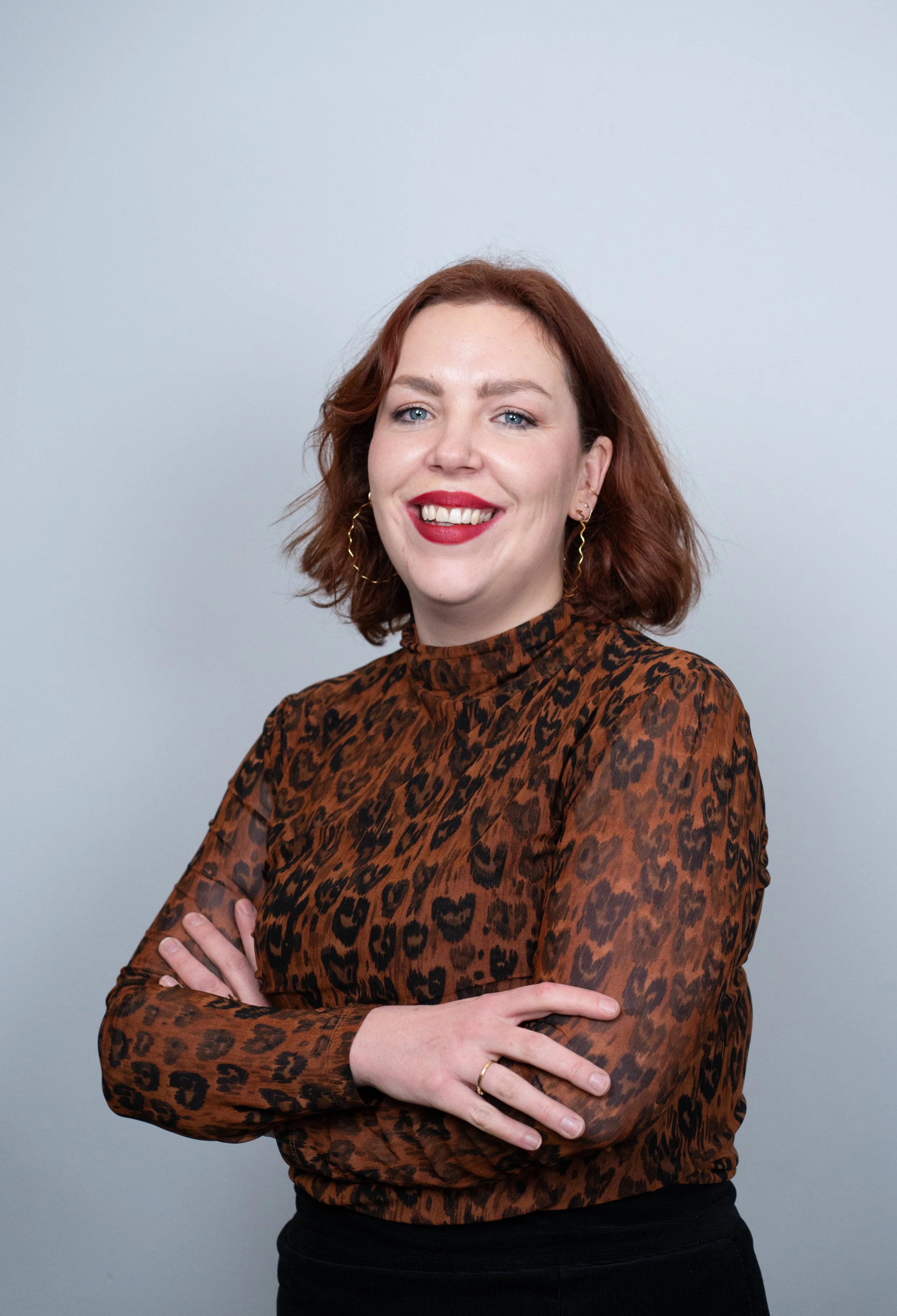 Portret van een lachende vrouw met rood haar, donkere leopardprint blouse en gouden oorbellen, staand met gekruiste armen voor een lichte achtergrond.