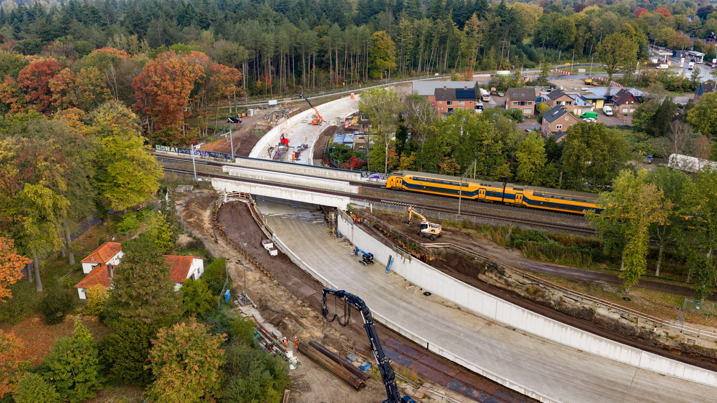 Een trein rijdt langs een bouwplaats voor een nieuwe weg of spoorlijn, omringd door bomen en huizen.