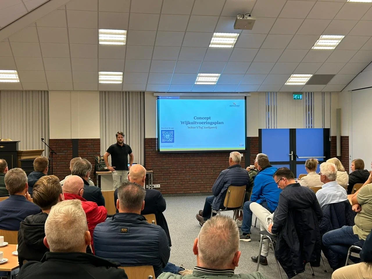 Een man geeft een presentatie aan een groep mensen in een vergaderzaal, met een scherm dat de tekst ‘Concept Wijkuitvoeringsplan’ toont.