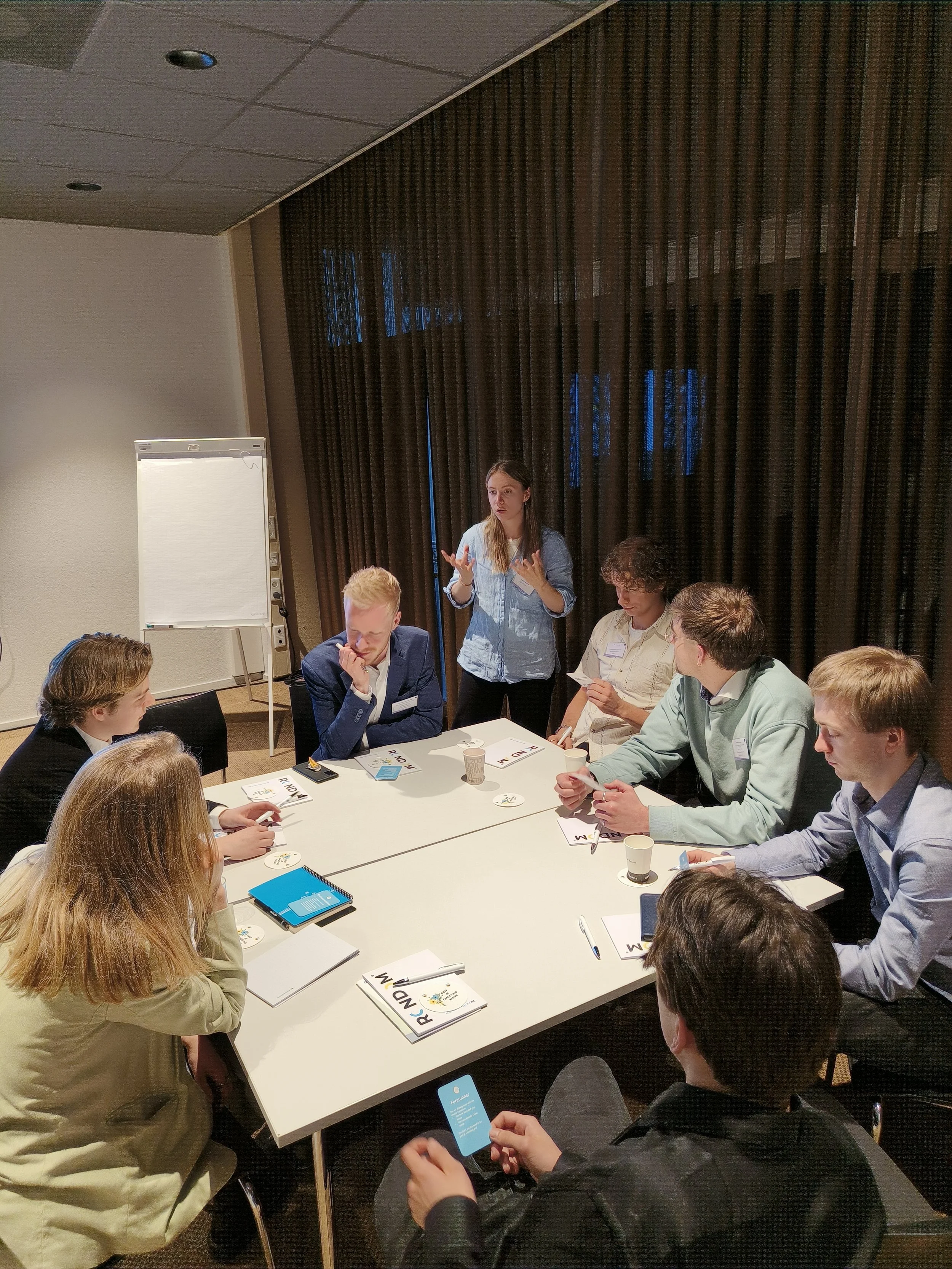 Een groep mensen zit aan een ronde tafel in een vergaderruimte, tijdens een presentatie of bespreking.