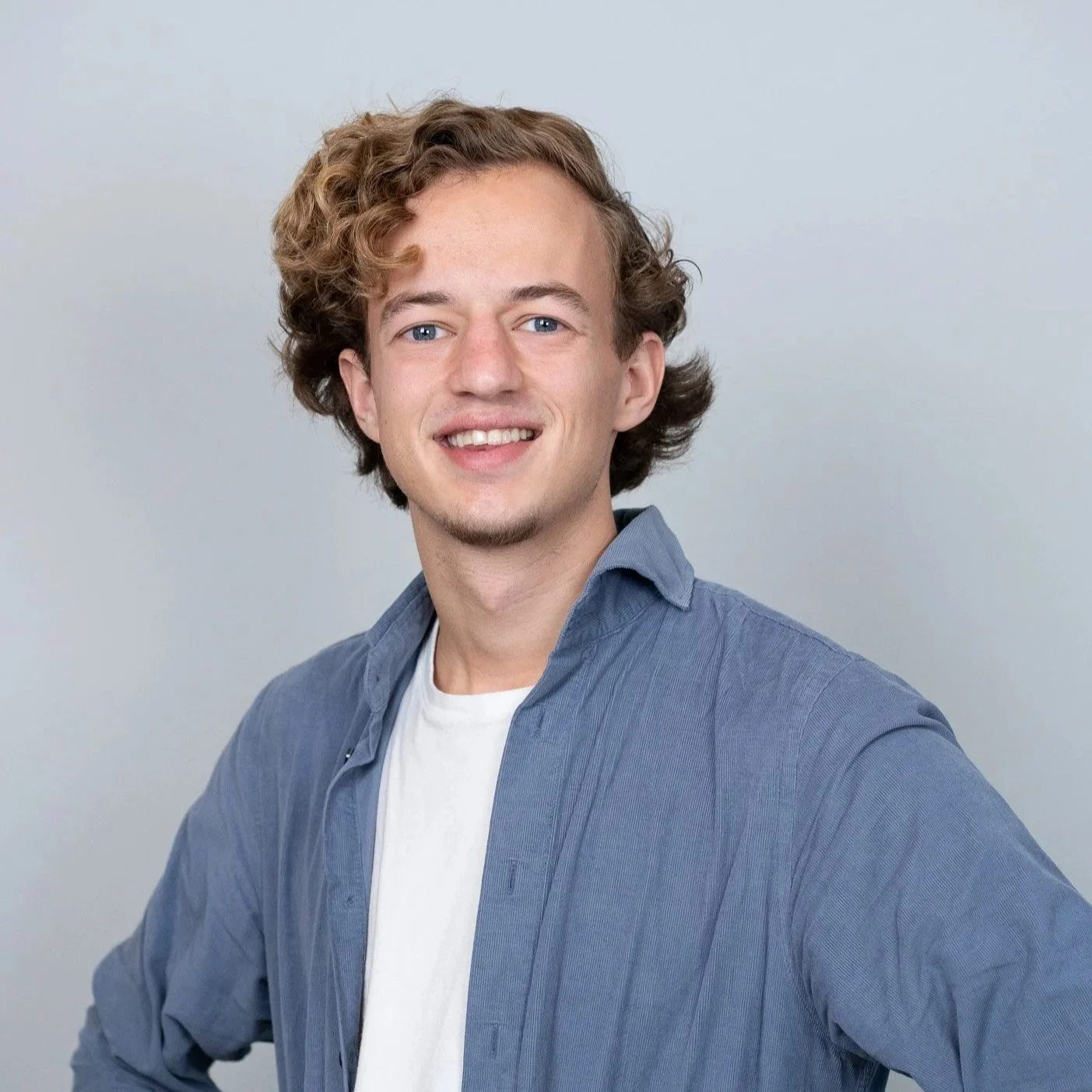 Portret van een jonge man met blond, gekruld haar, blauwe ogen, lichte huid, chatten, lachend, blauwe overhemd over een witte t-shirt, op een lichte achtergrond.