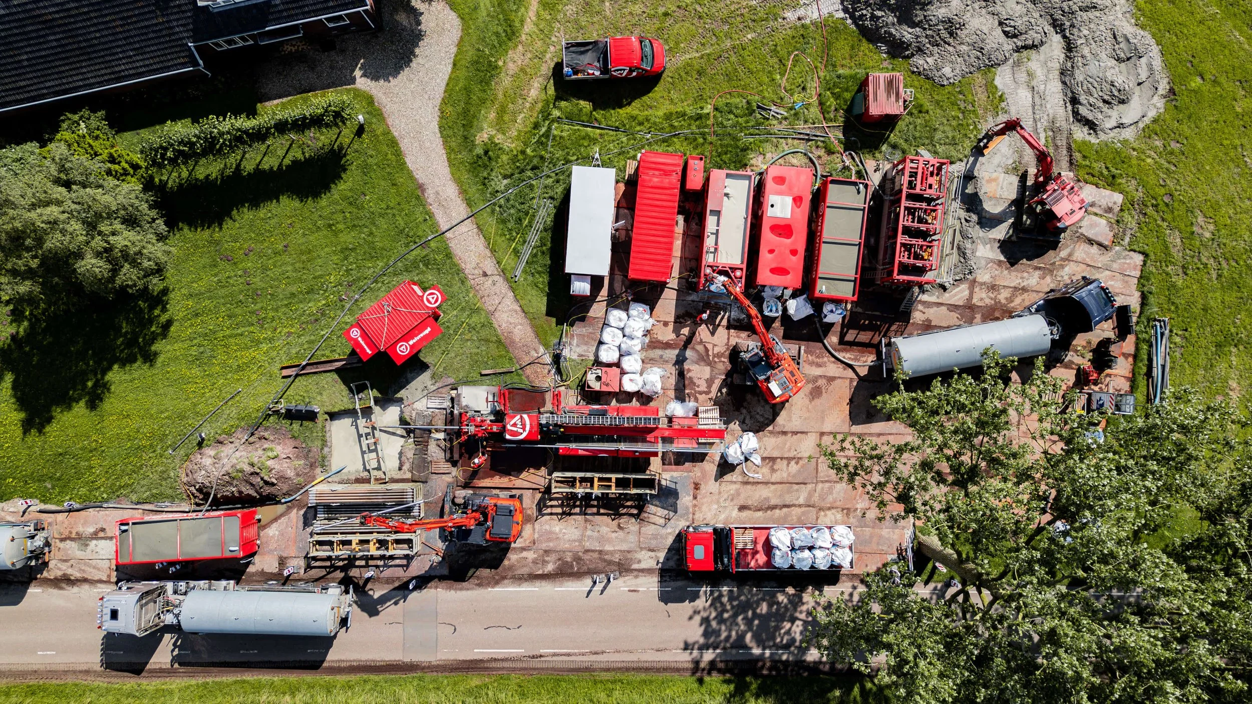 Bovenaanzicht van een bouwplaats met grote rode machines, opslagcontainers, en bouwmaterialen, naast een weg en groene grasvelden. 
