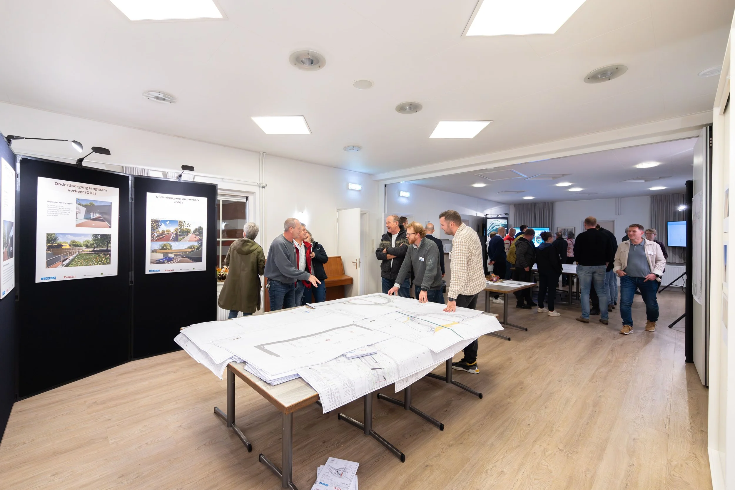 Informatieve bijeenkomst met mensen die rond een tafel met bouwplannen zitten, posters over verkeersprojecten en een groep mensen in een ruime, goed verlichte kamer.
